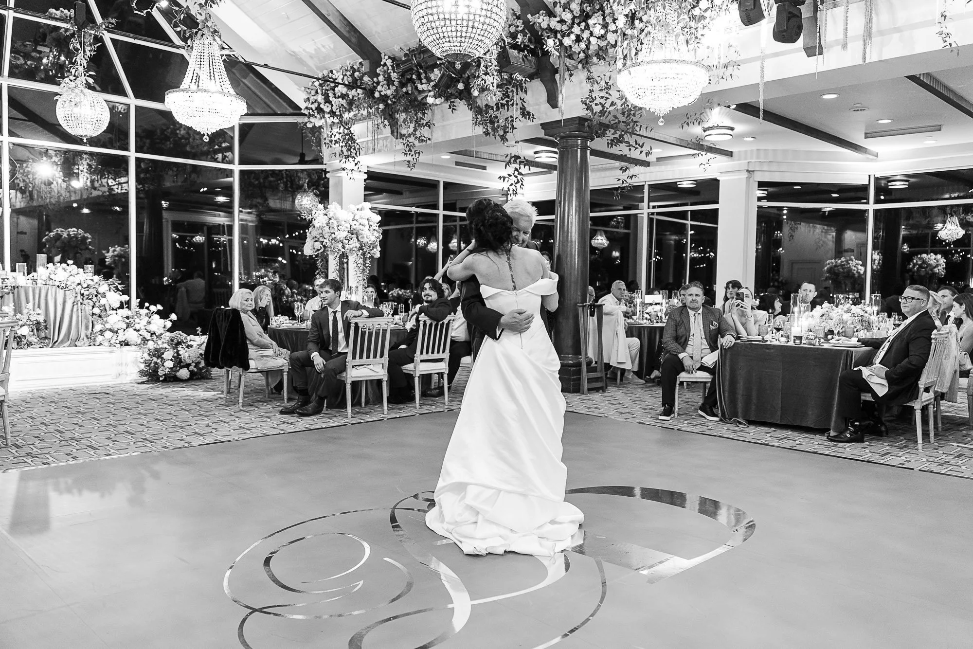 A bride and groom sharing a dance at their wedding reception, with guests seated at decorated tables watching in a glass-walled reception hall decorated with flowers and hanging chandeliers at the Beach and Tennis Club in Pebble Beach, CA.