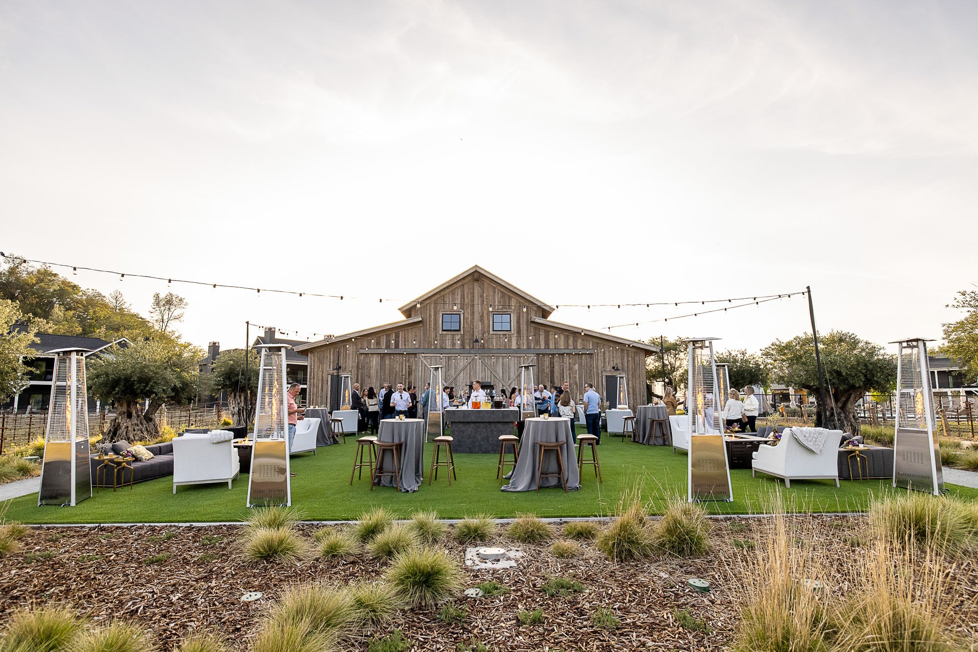 Outdoor event reception at sunset with guests mingling, standing tables, lounge seating, and patio heaters in front of a rustic barn-style building adorned with string lights at the Napa Valley Four Seasons Hotel.
