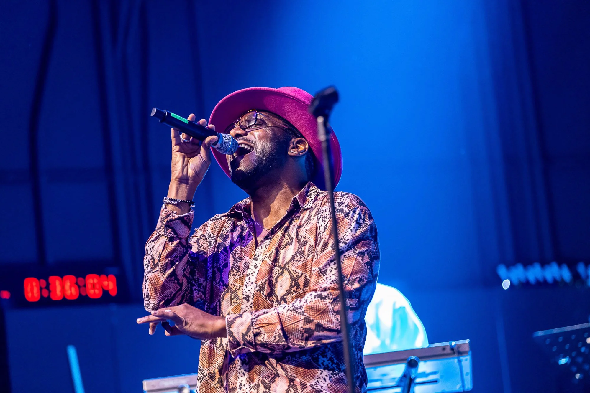 Wyclef Jean wearing a pink hat, glasses, and a snake-print shirt singing into a microphone on stage at the Fox Theater in Oakland.