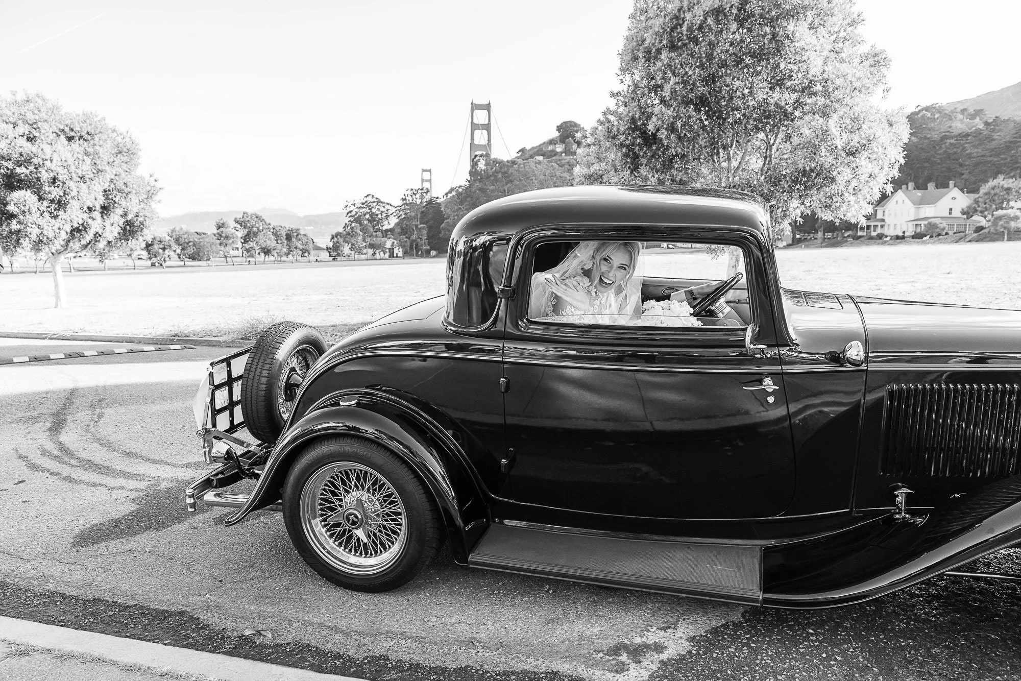 Black vintage car with a smiling woman inside, in an outdoor park setting, with trees and hills in the background at Cavallo Point in Sausalito, CA.