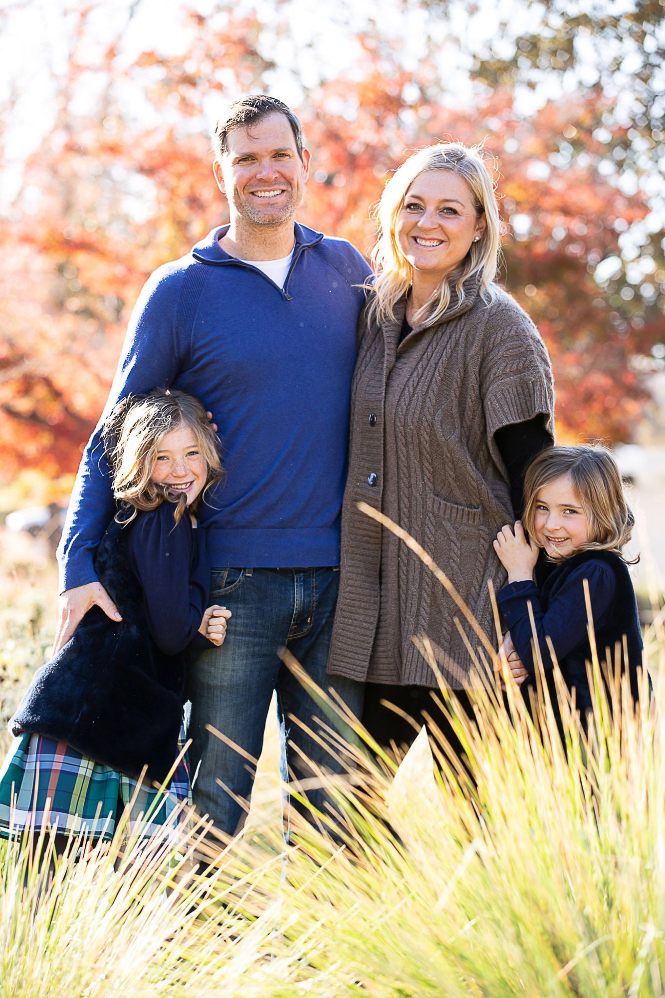 A happy family of four, including a man, woman, and two young girls, standing outdoors on a sunny day surrounded by autumn foliage, smiling at the camera.