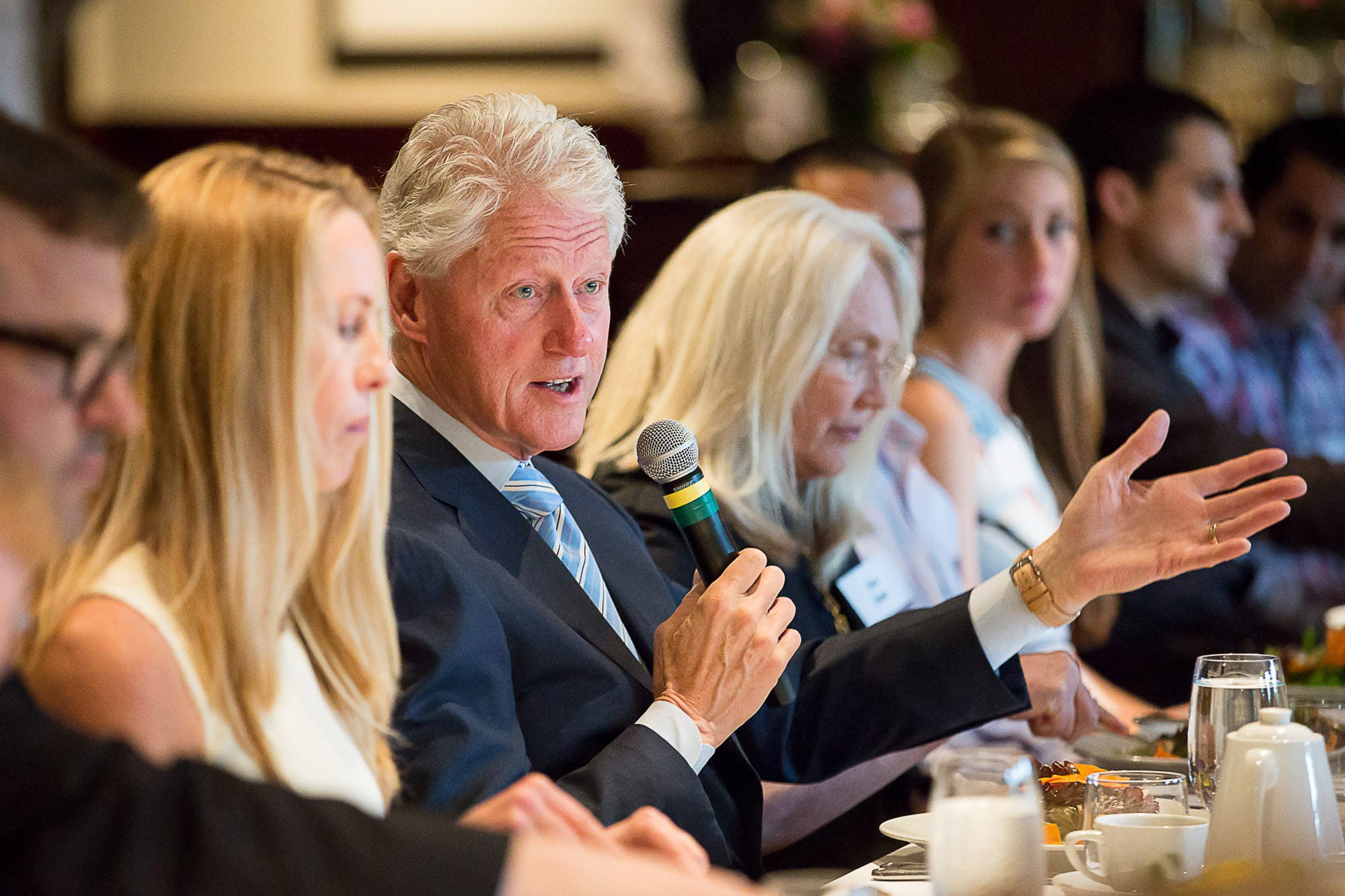Former President Bill Clinton with gray hair and a suit speaking into a microphone during a panel discussion at a formal event, seated among other women and men.