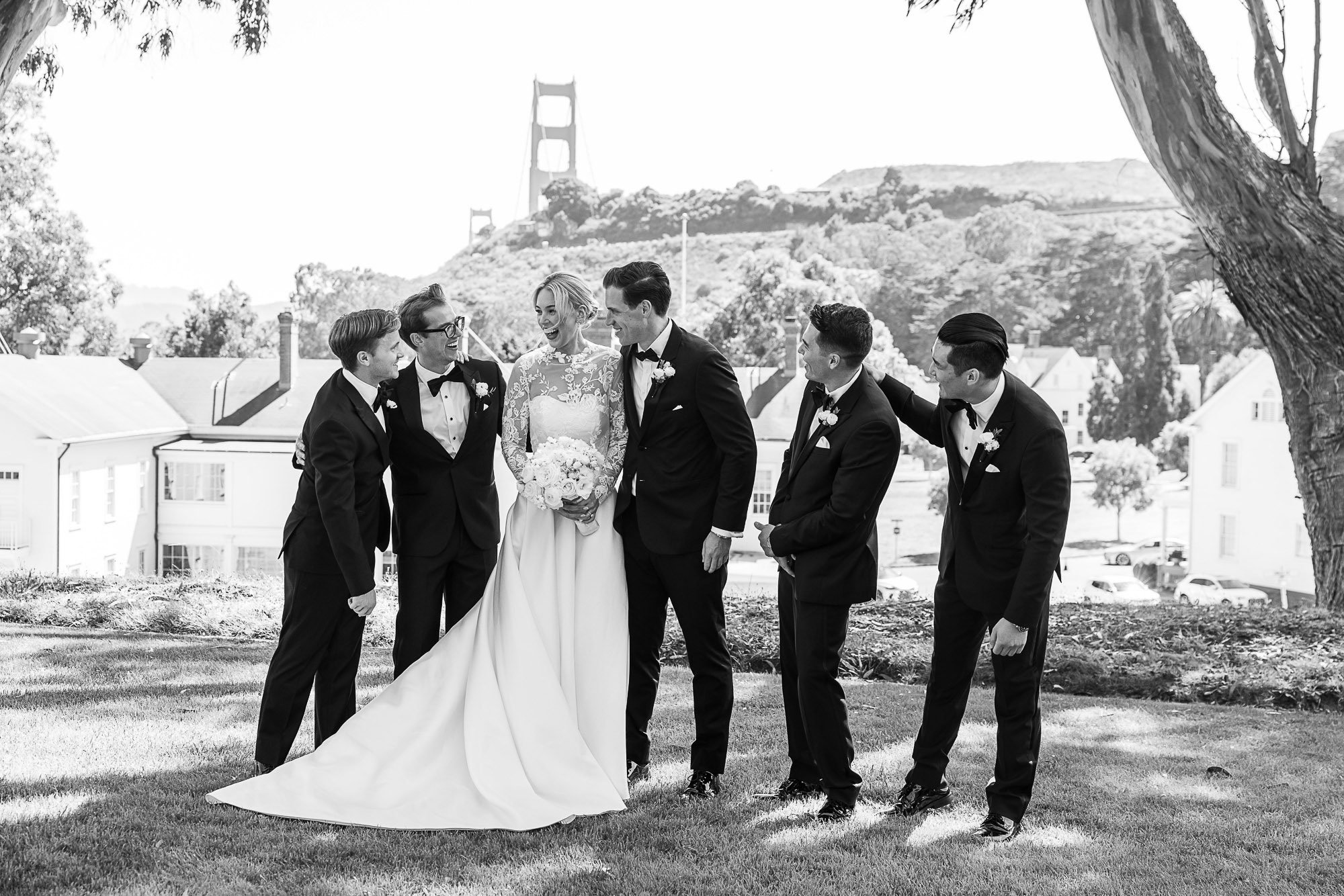 Black and white photo of a bride in a wedding dress and six groomsmen in tuxedos standing outdoors near trees with the Golden Gate Bridge visible in the background at Cavallo Point in Sausalito, CA.