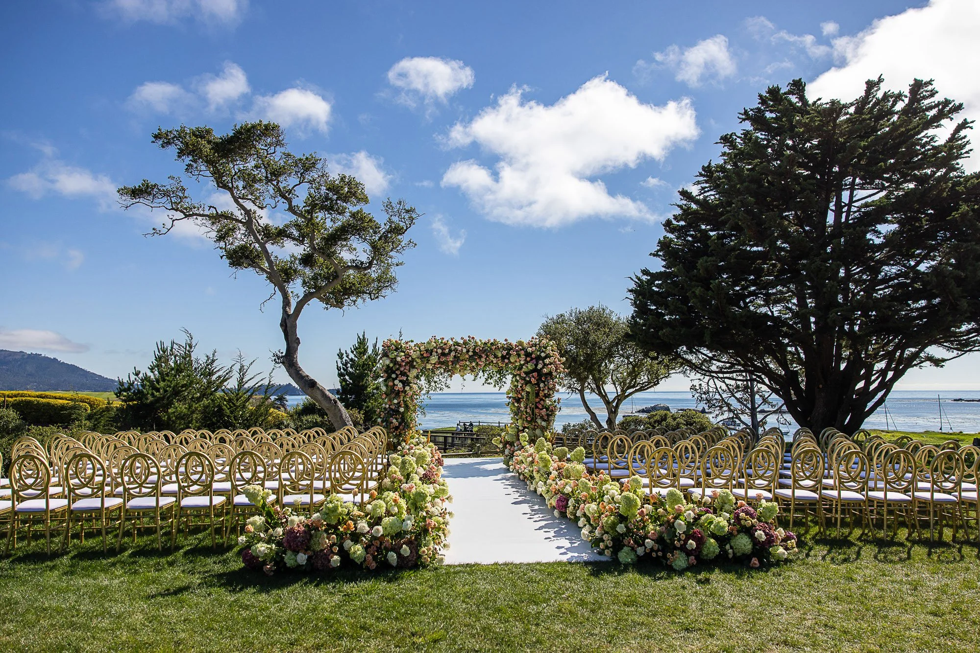 Outdoor wedding setup with rows of chairs facing a floral arch overlooking the water, surrounded by trees and blue sky at the Beach and Tennis Club in Pebble Beach, CA.