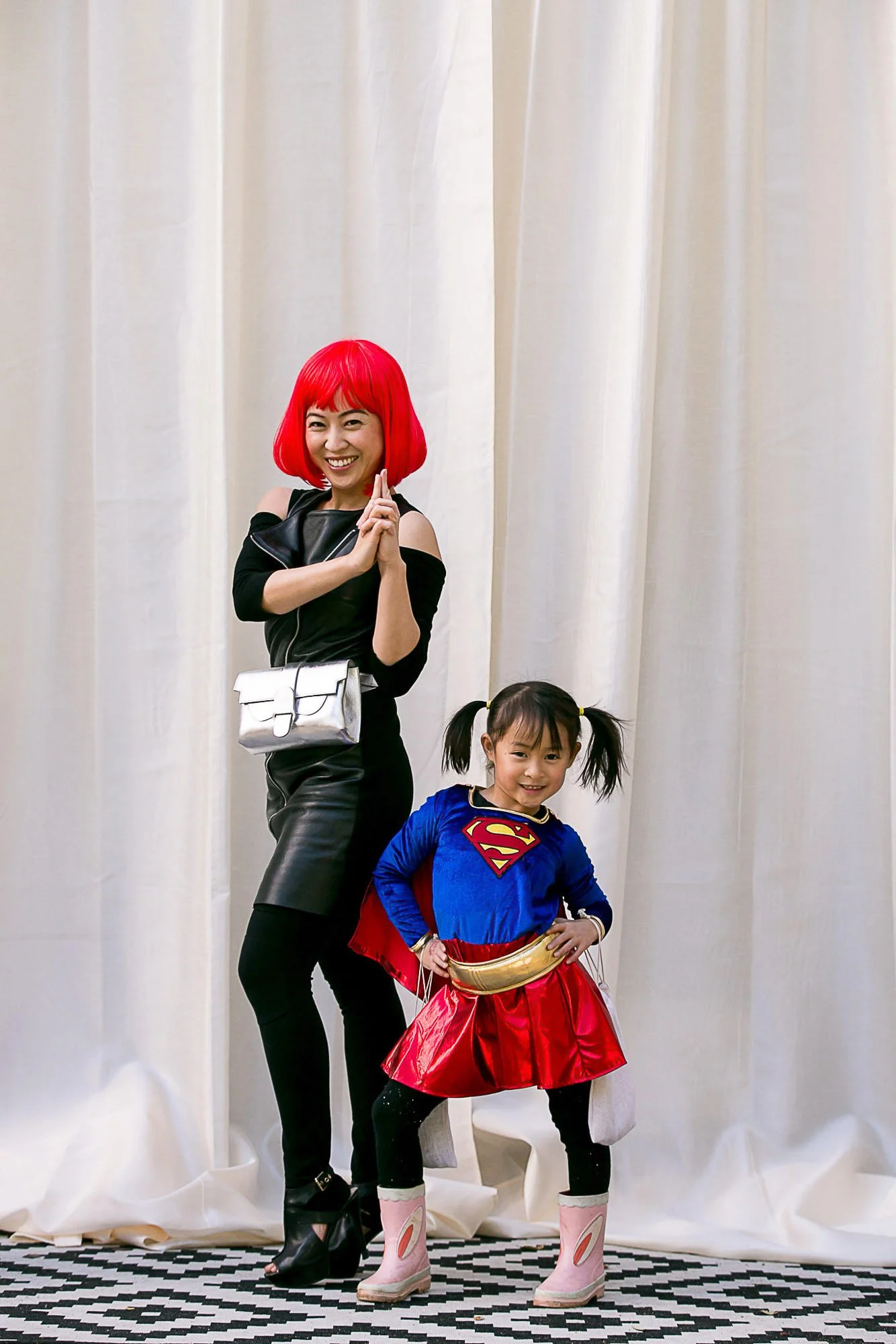 A woman with bright red hair and a young girl dressed as Supergirl, both smiling and posing indoors in front of a cream-colored curtain.