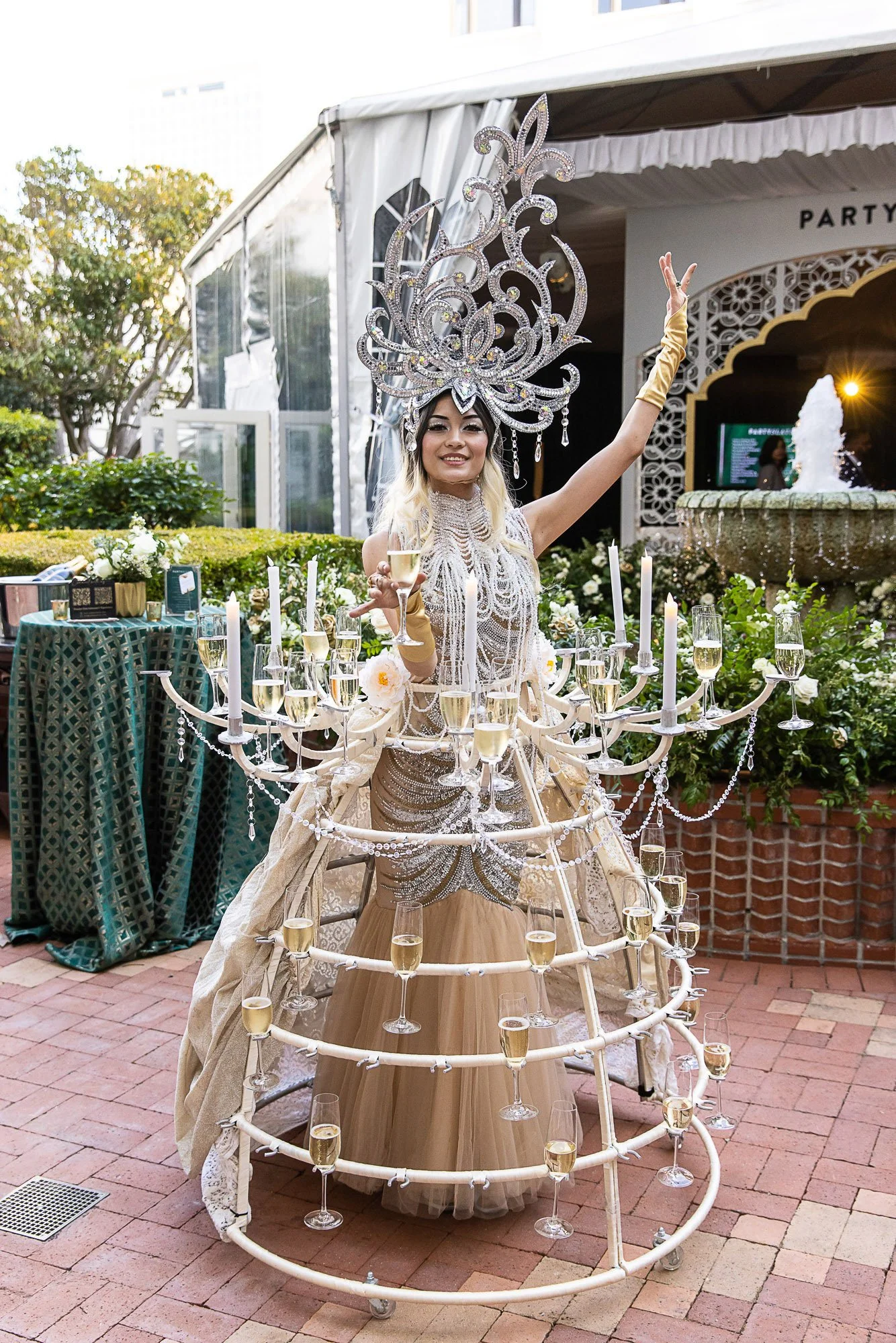 Woman in elaborate costume with large silver headdress, holding a champagne glass, surrounded by candles and champagne flutes, outdoors in a garden event setting.