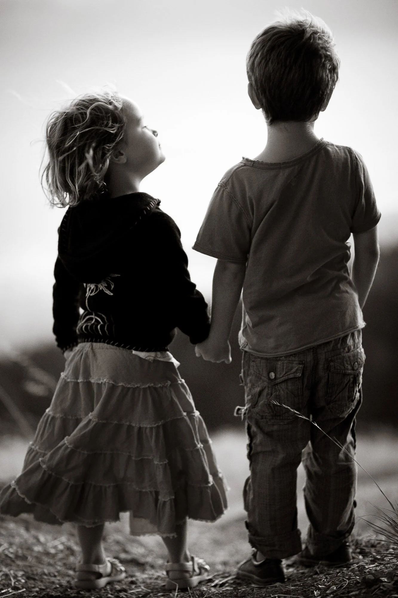 Two children holding hands, walking outdoors at sunset or dusk, with their backs to the camera; girl wearing a layered ruffled skirt and girl wearing shorts and a t-shirt, both in a black and white photo.