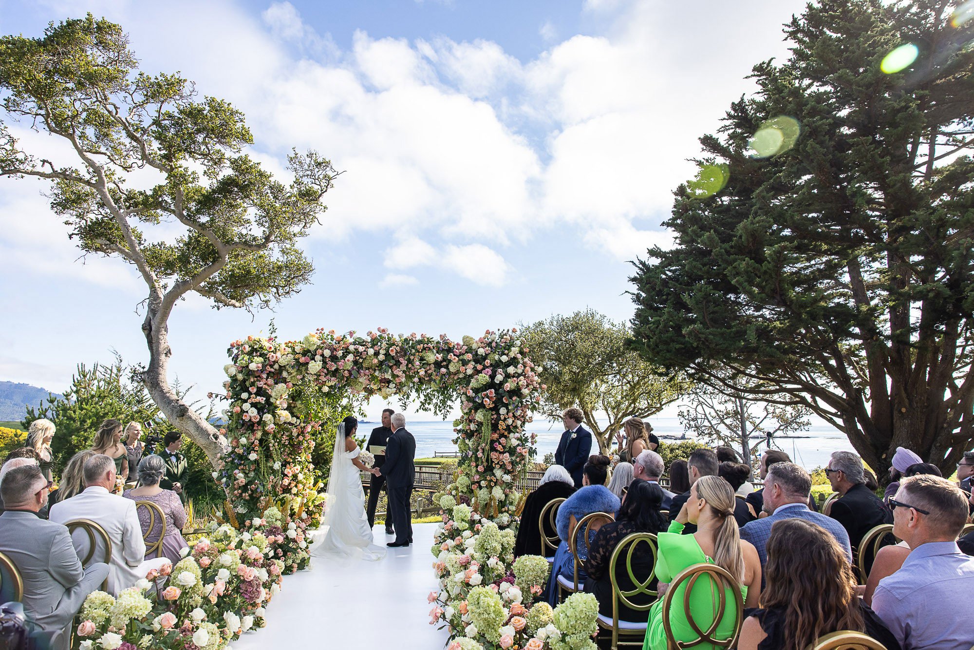 Outdoor wedding ceremony at the Beach and Tennis Club in Pebble Beach with a floral arch, guests seated on either side, and trees under a partly cloudy sky.