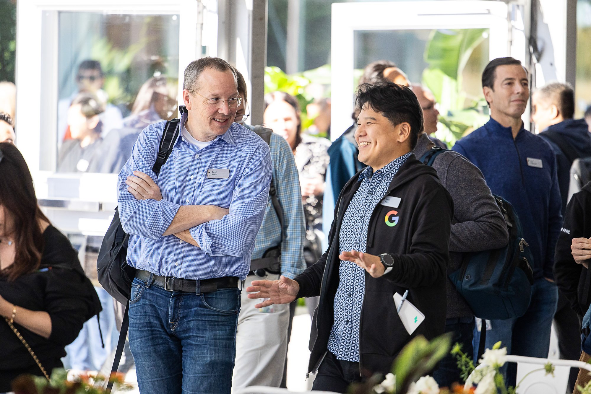 Two men engaged in conversation at a professional event, with other attendees in the background, and greenery visible outside through large windows.
