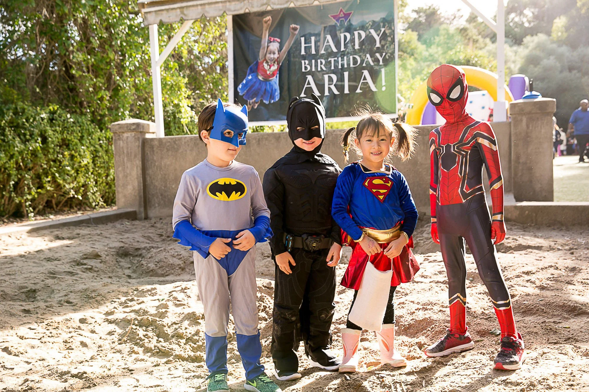 Four children dressed in superhero costumes standing outdoors on a sunny day in front of a sign that says 'Happy Birthday Aria!'. The children are dressed as Batman, Black Panther, Supergirl, and Spiderman. There are trees and a playground in the bac