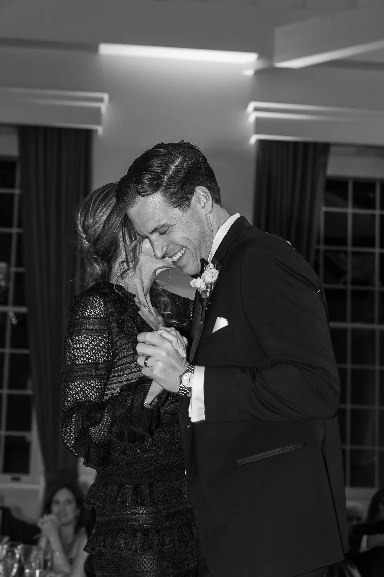 A black-and-white photo of a joyful man and woman dancing closely together, smiling, in an elegant indoor setting. They are holding hands, with the woman's head resting against the man's forehead. The man is wearing a tuxedo with a bow tie and a bout