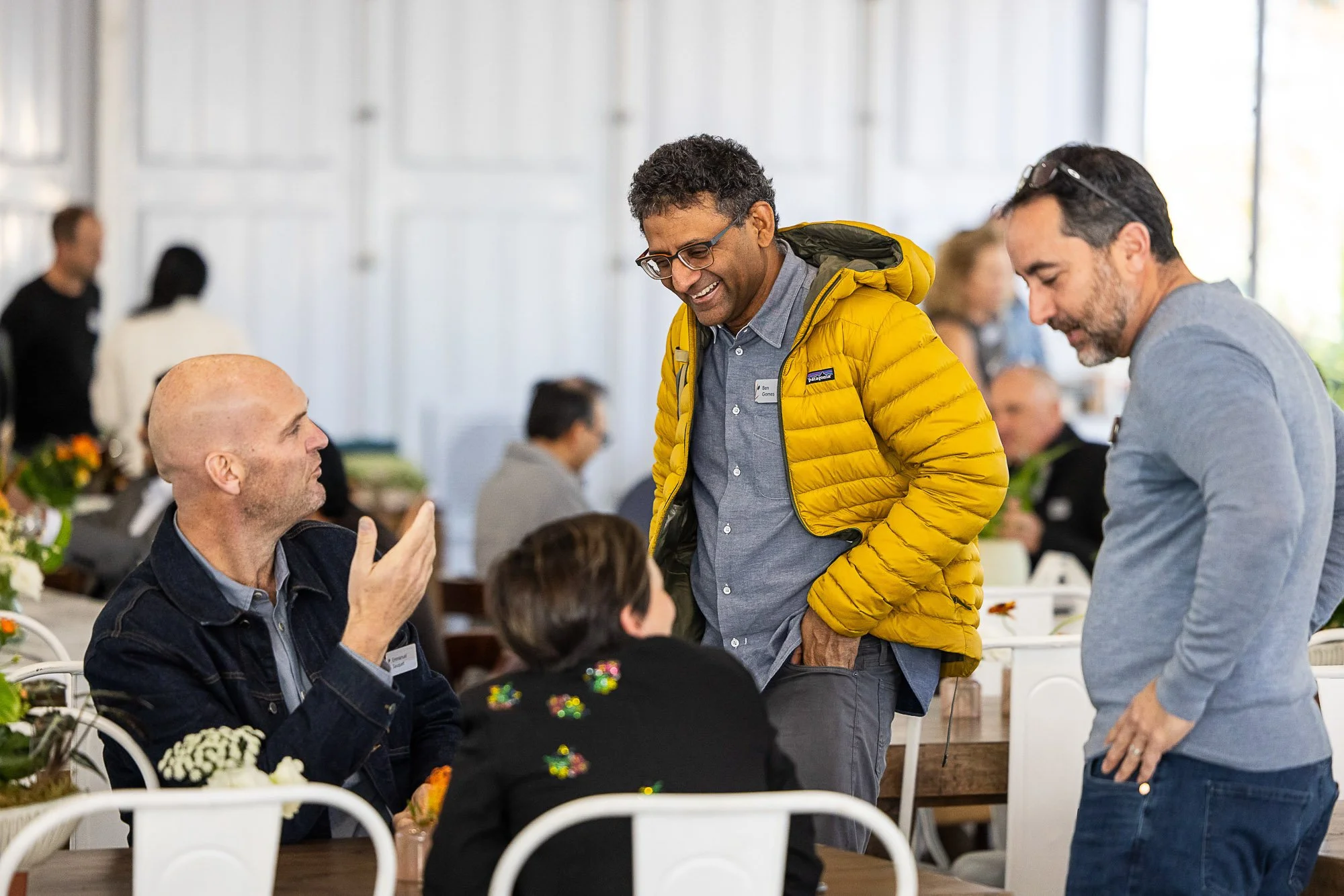 People at a social gathering or event, seated at a table with white chairs, in a bright indoor space with large windows. Two men are standing and talking, one wearing a yellow jacket and the other in a gray shirt, while a bald man with a beard and a 