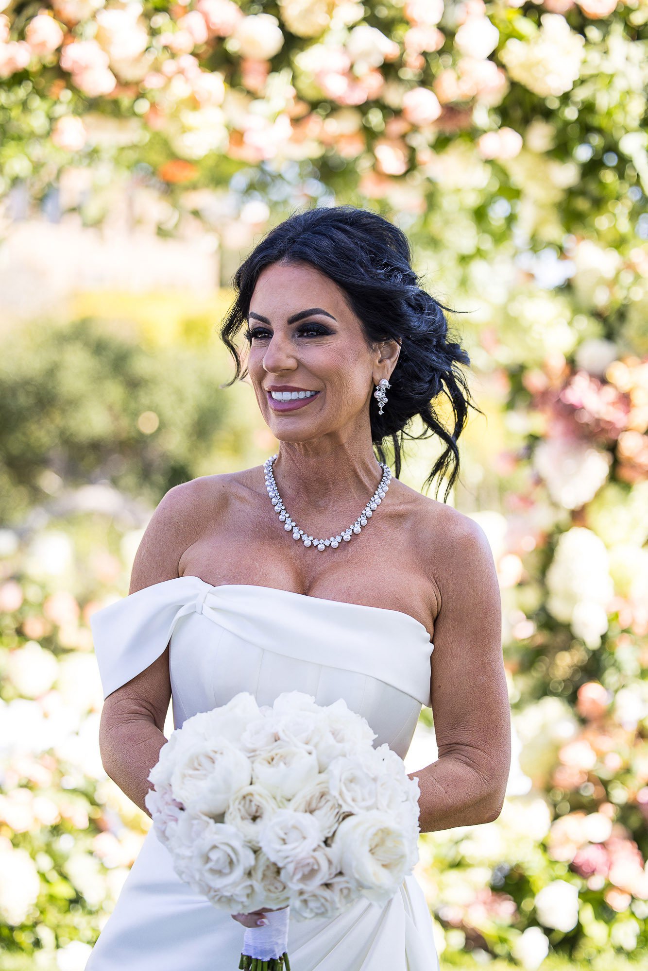 A bride with dark, curly hair in an updo, wearing a strapless white wedding gown with a bow detail, holding a bouquet of white roses, and standing outdoors at the Beach and Tennis Club in Pebble Beach, CA.