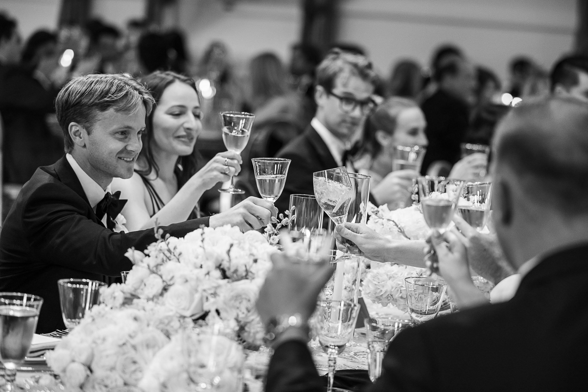Black and white photo of wedding reception with guests raising glasses for toast at a decorated table with flowers at Cavallo Point in Sausalito, CA.