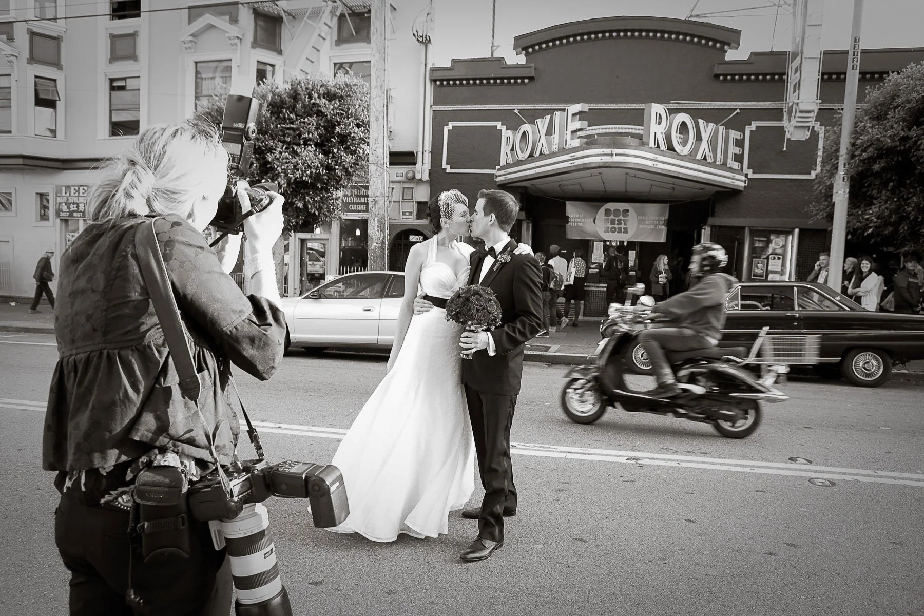 A bride and groom kissing in front of the Roxie Theater in San Francisco, with photographer Tara Arrowood taking their picture on the street. A motorcycle rides by, and pedestrians are visible in the background.