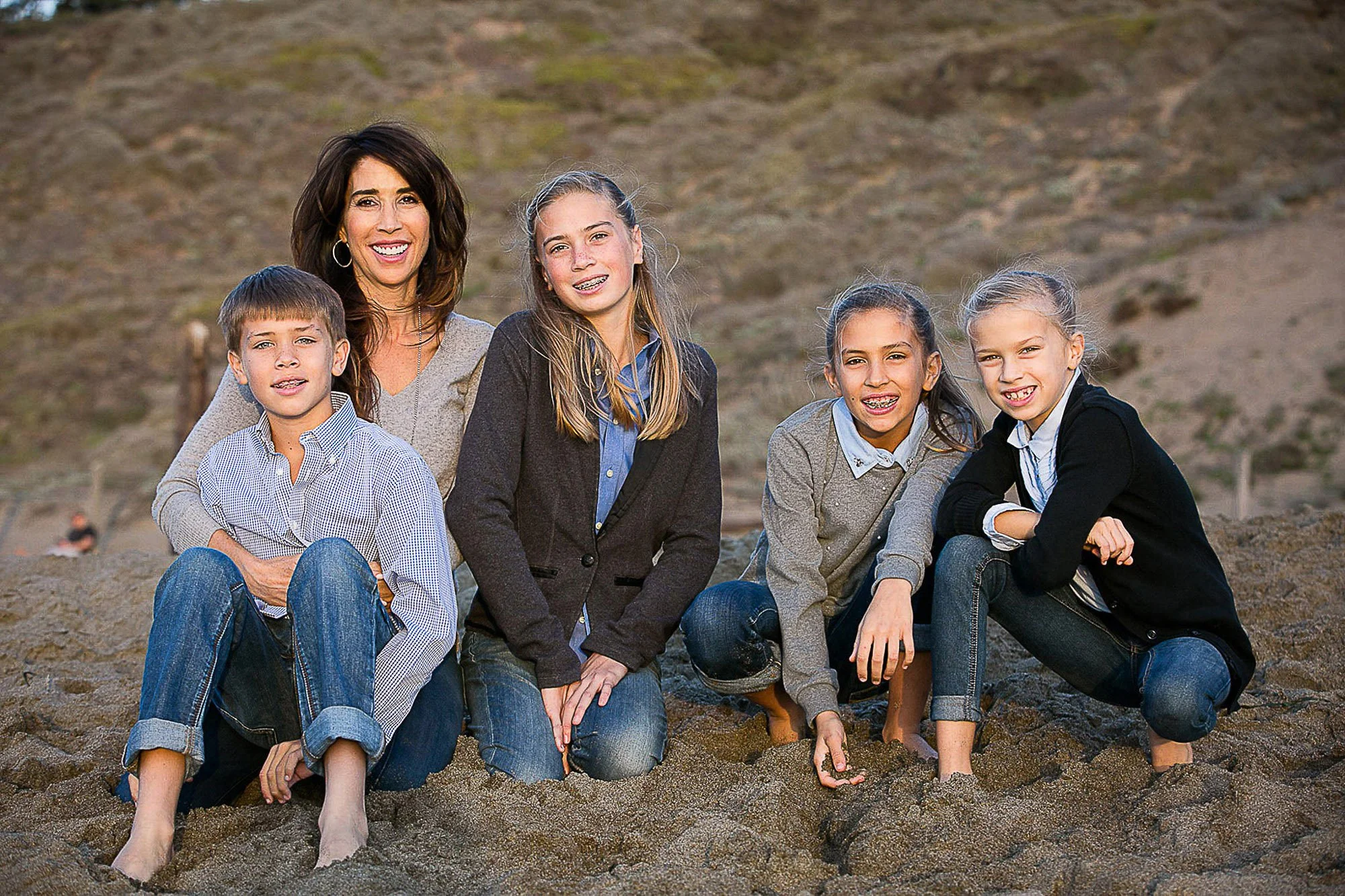 A group of five people, one adult woman and four children, sitting on a sandy beach with hills in the background. They are smiling and looking at the camera at Ocean Beach in San Francisco California.