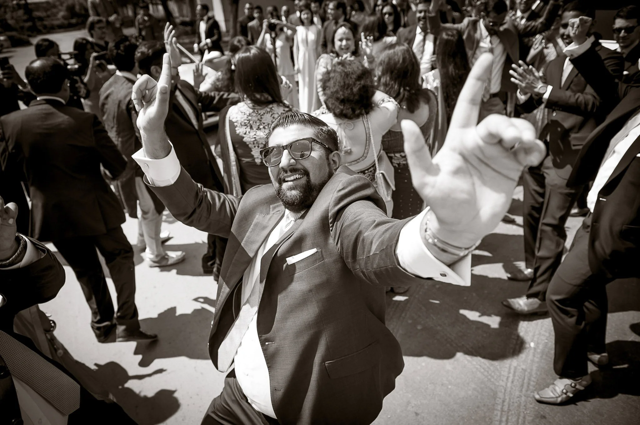 A group of people, dressed in formal and casual attire, gathered outdoors and dancing to music. A man in front wearing sunglasses and a suit is smiling and pointing towards the camera.