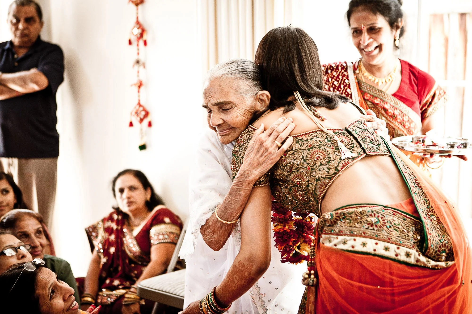 An elderly woman dressed in traditional Indian attire hugging a young woman during a celebration. The scene is festive with other women in colorful clothing watching and smiling.