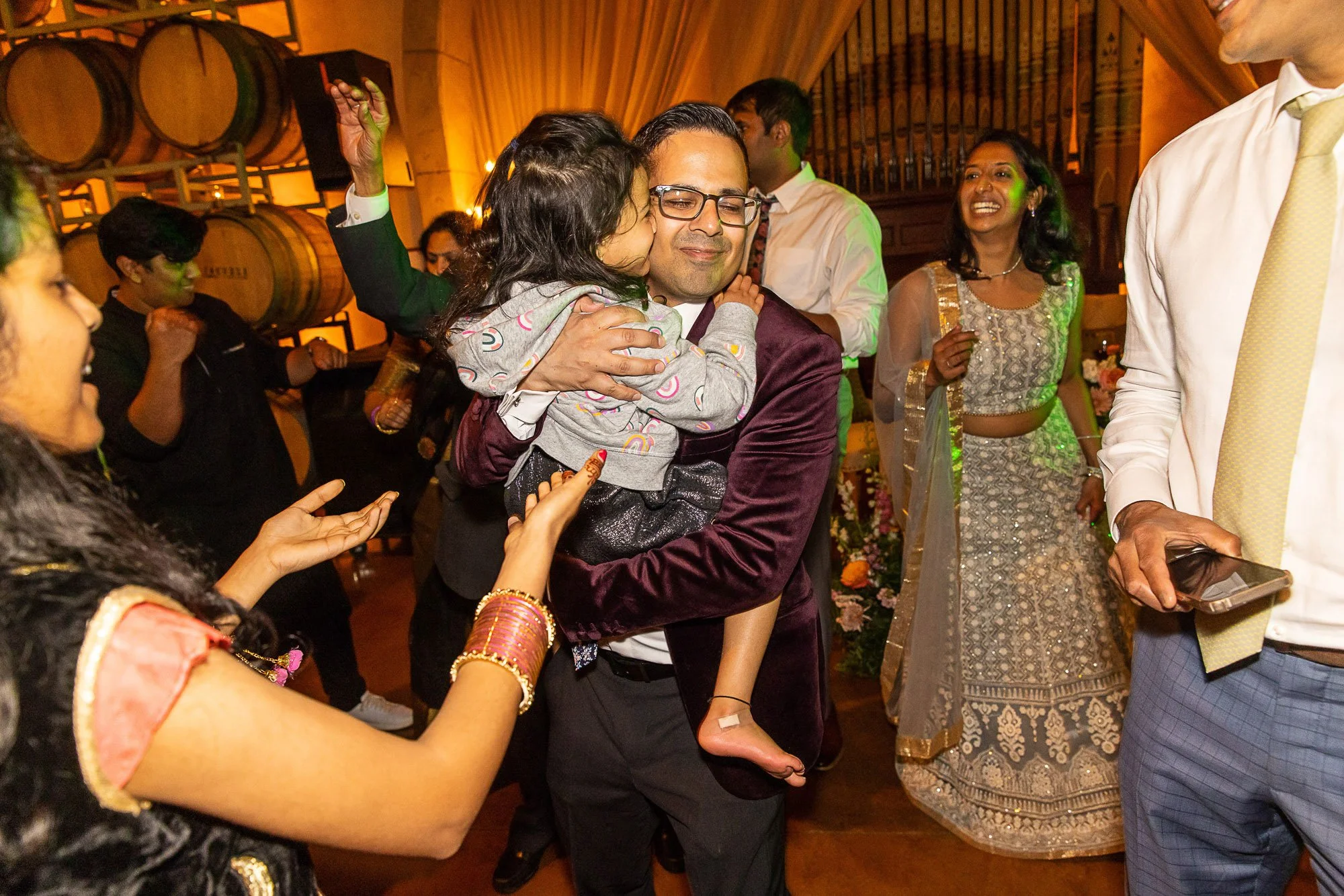 People celebrating at a wedding reception, with a woman kissing a man on the cheek, the man smiling and wearing glasses, surrounded by other guests in traditional and formal attire in a decorated indoor venue with wooden barrels and warm lighting.