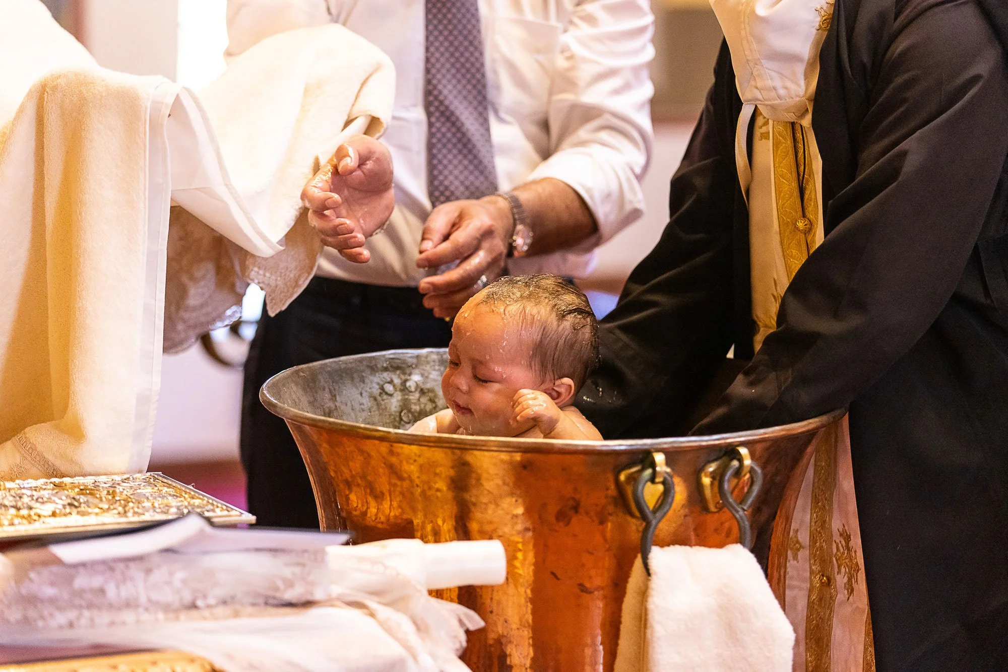 A baby is getting baptized in a metal bowl during a religious ceremony, with several people gathered around.
