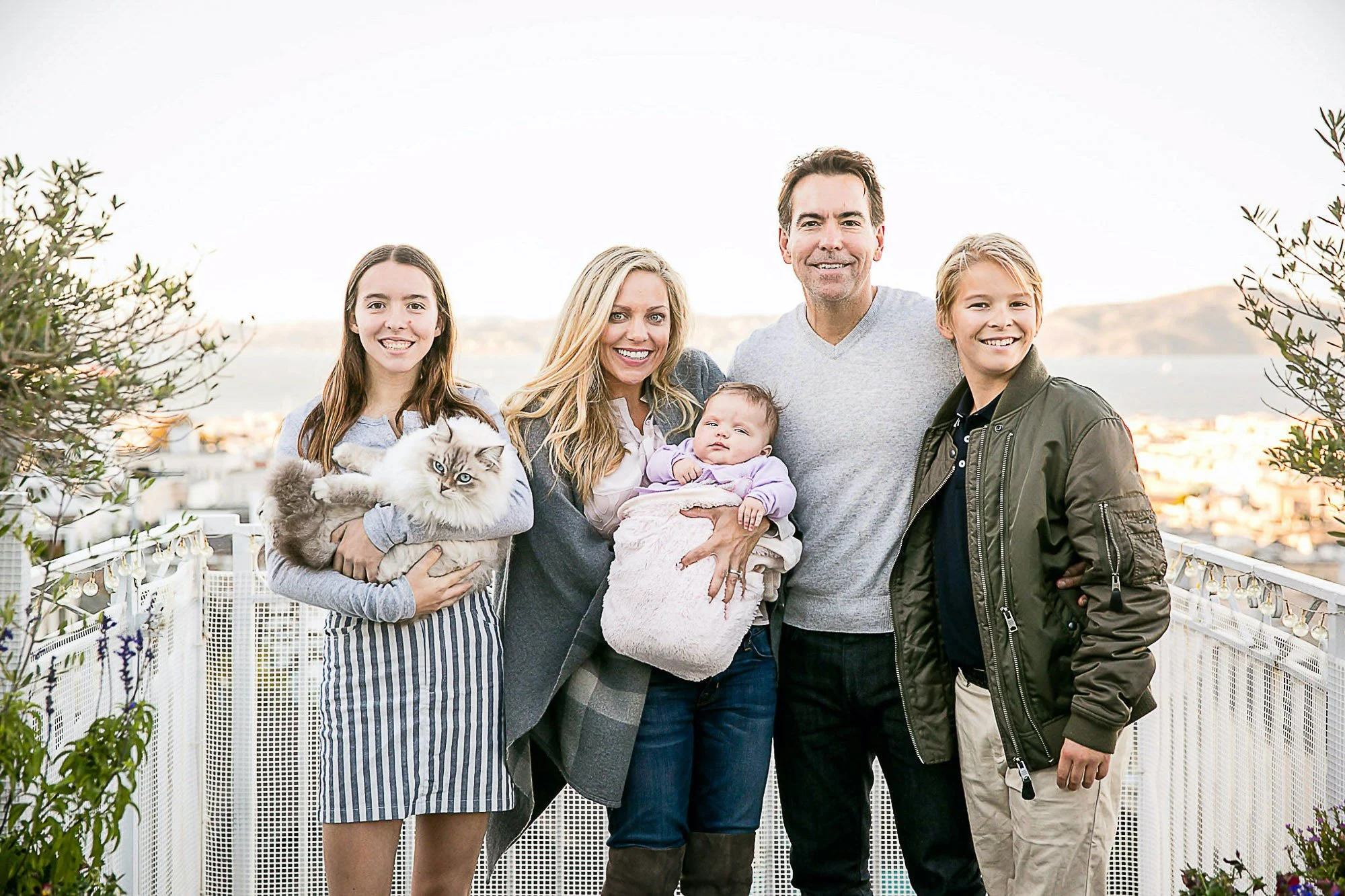 A group of six people, including two young girls, two adults, a baby, and a teenage boy, standing together outdoors on a balcony with plants and a cityscape in the background. One of the girls is holding a fluffy gray and white cat.
