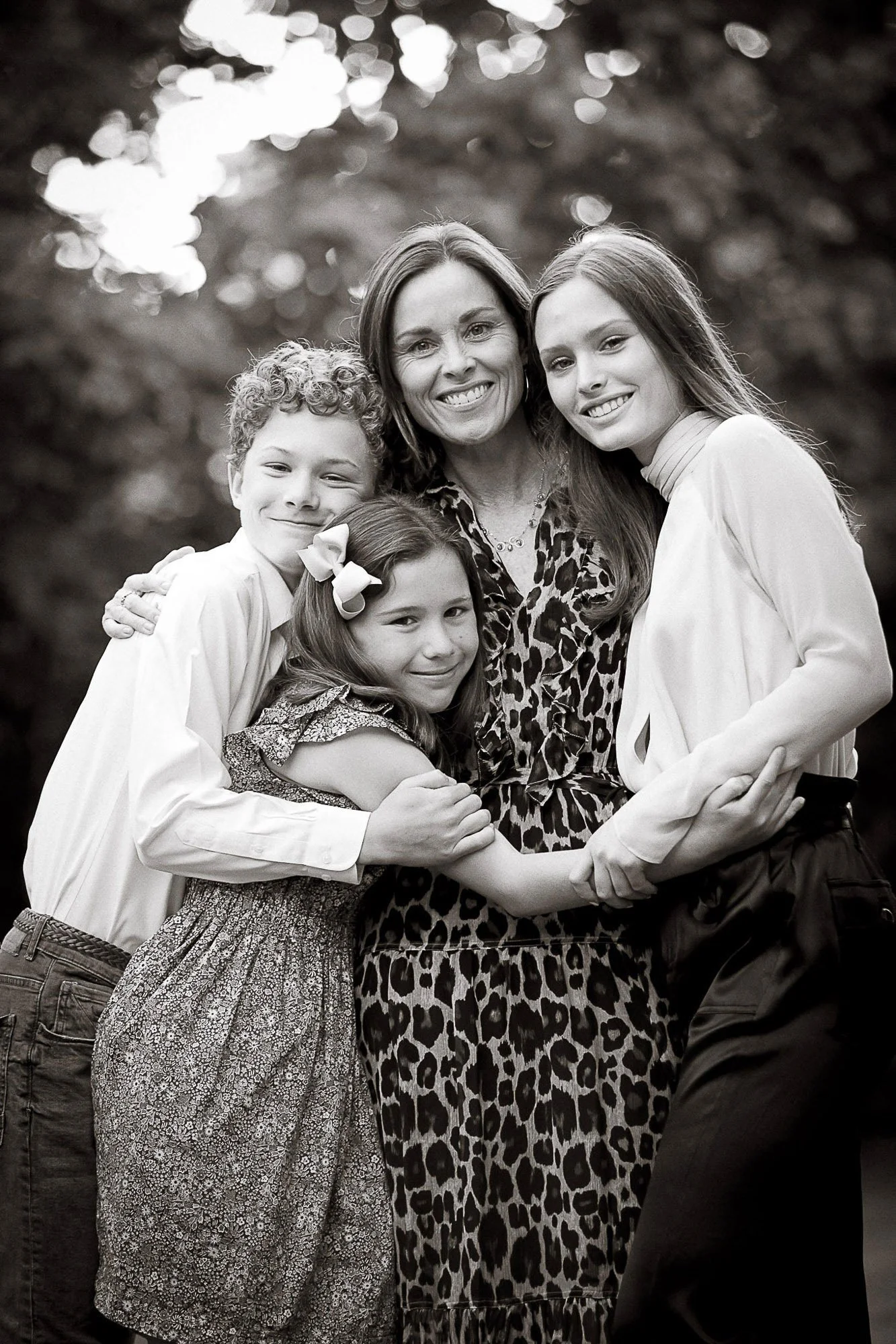 Black and white photo of five smiling people, including an adult woman and four children, hugging each other outdoors with a blurred leafy background.