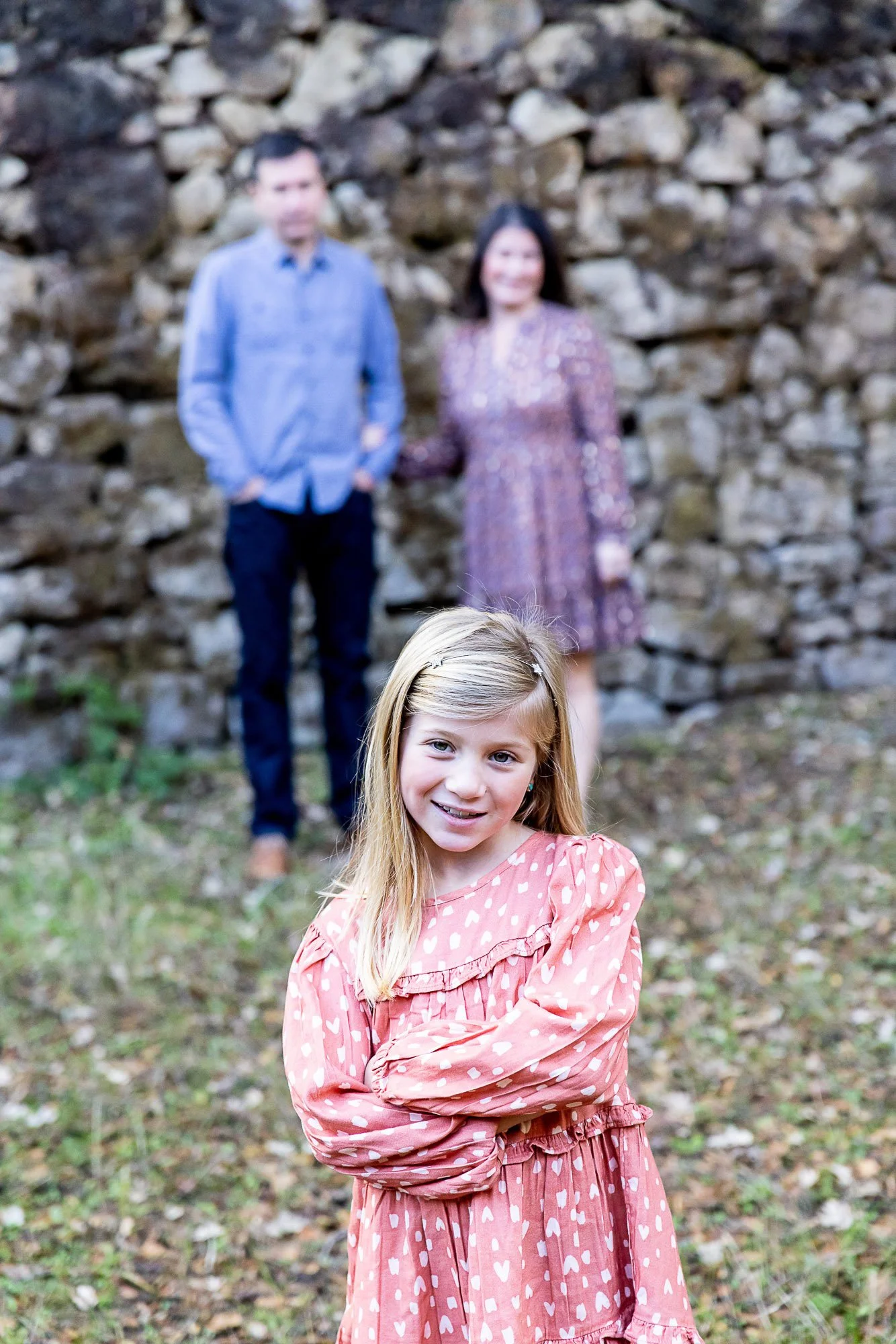 A young girl with blonde hair smiling, standing outdoors in a pink dress with white hearts, in front of a stone wall, with a man and woman in the background.