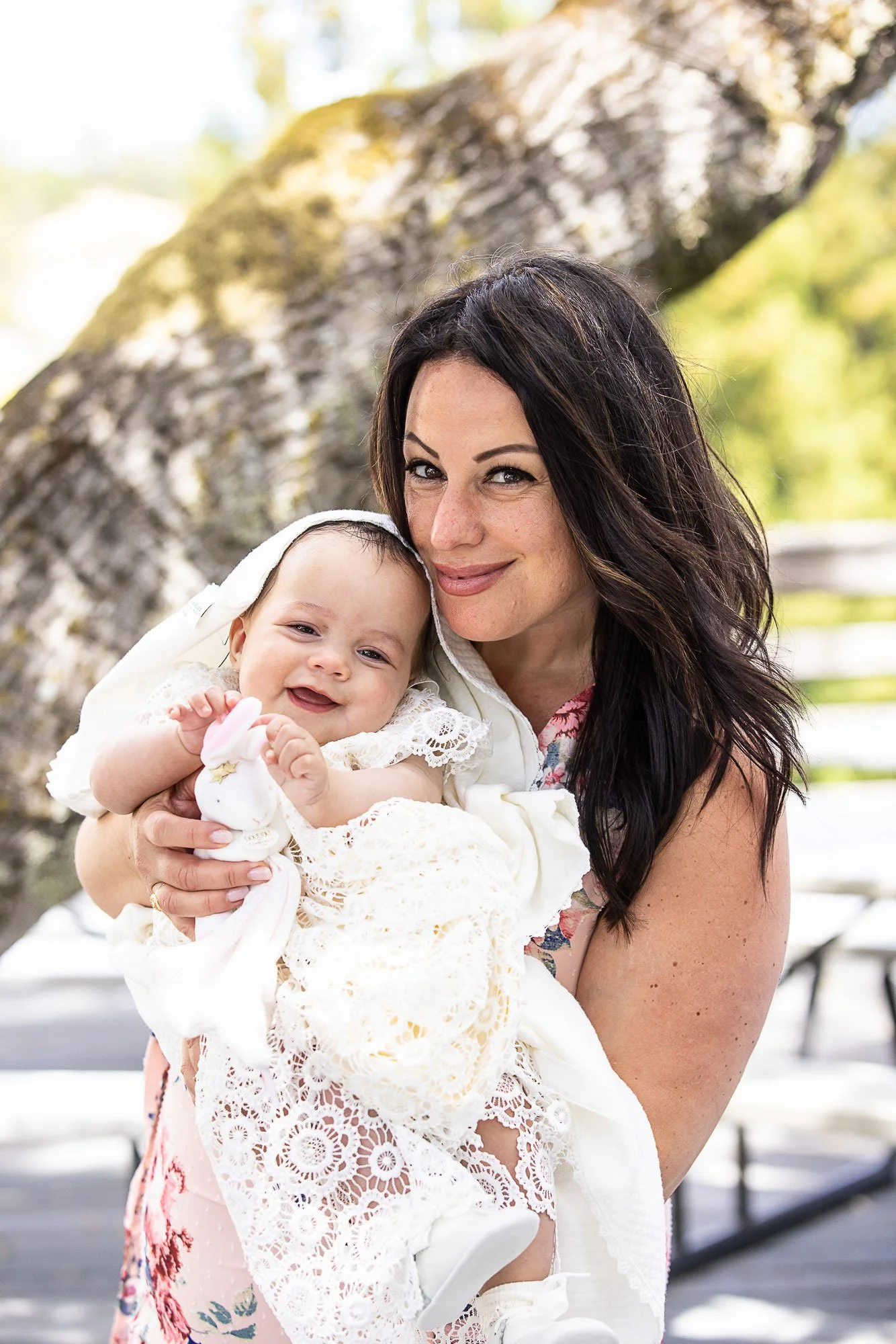 A woman with dark hair holding a smiling baby girl with a white bonnet and dress outdoors with trees and a bench in the background.