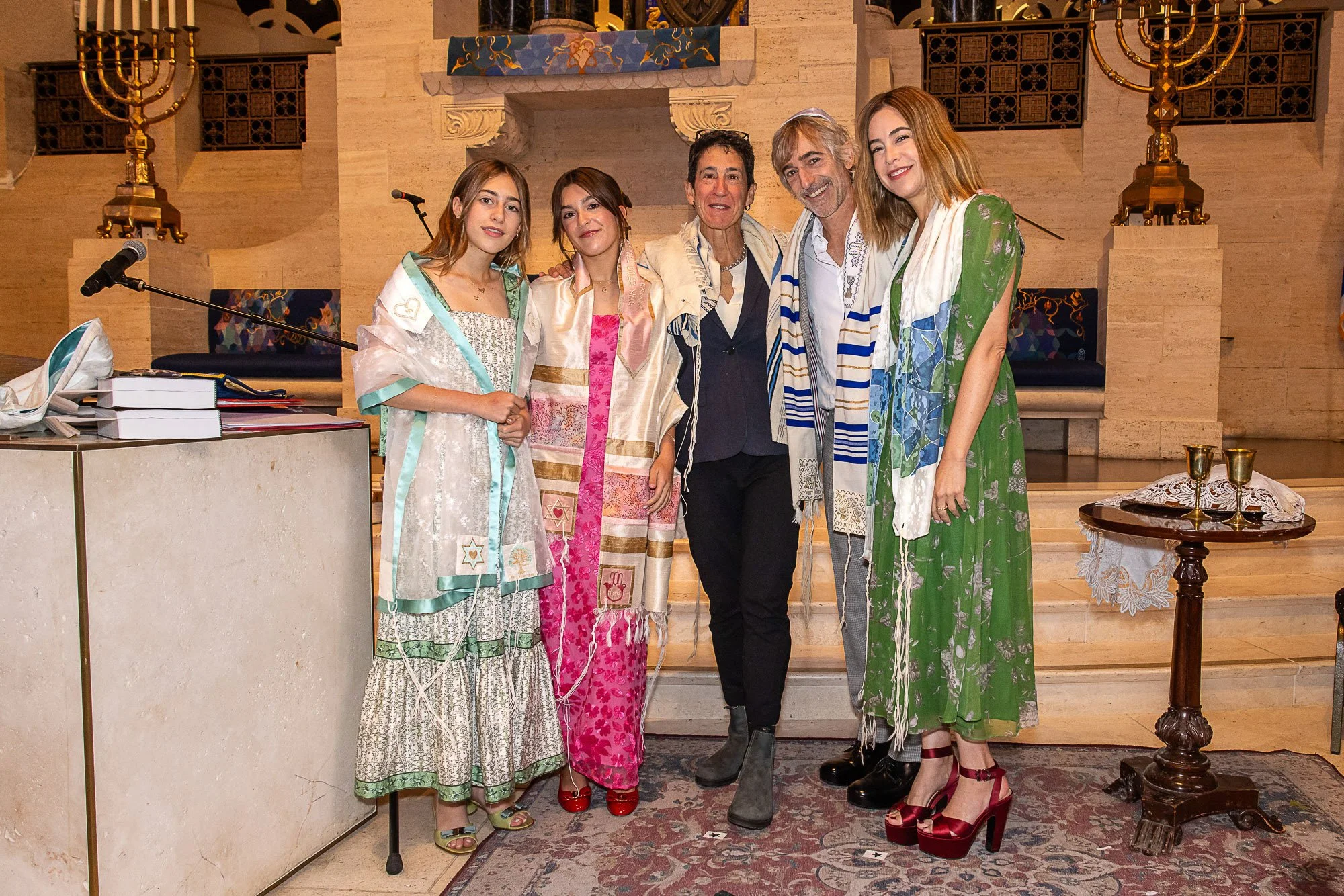Group of five people, four women and one man, standing together inside a building with religious symbols and menorahs, smiling and posing for the photo at a Bat Mitzvah.
