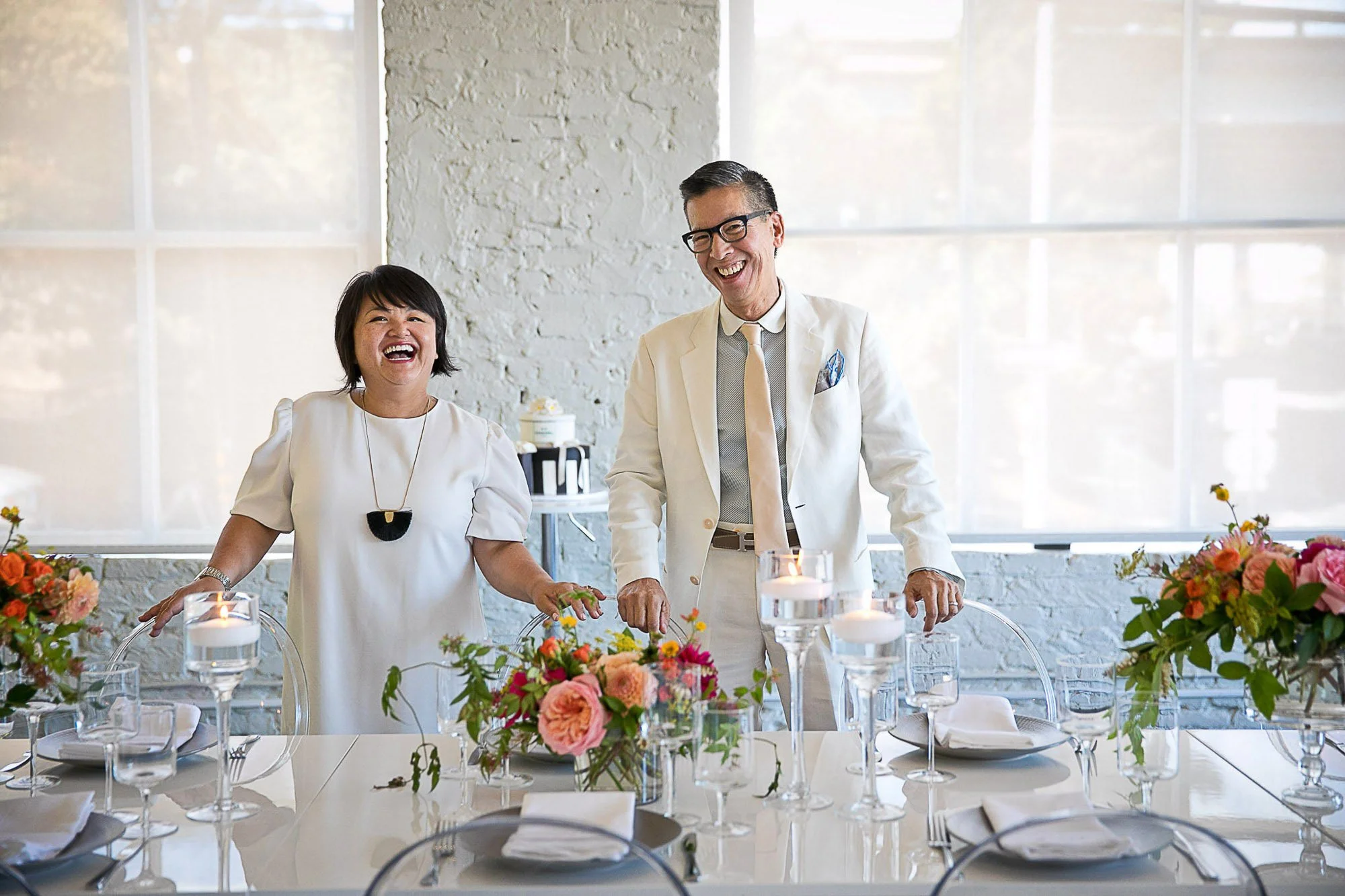 Two people smiling and laughing at a decorated table with candles and flowers, likely at a celebration or event, inside a bright room with large windows.