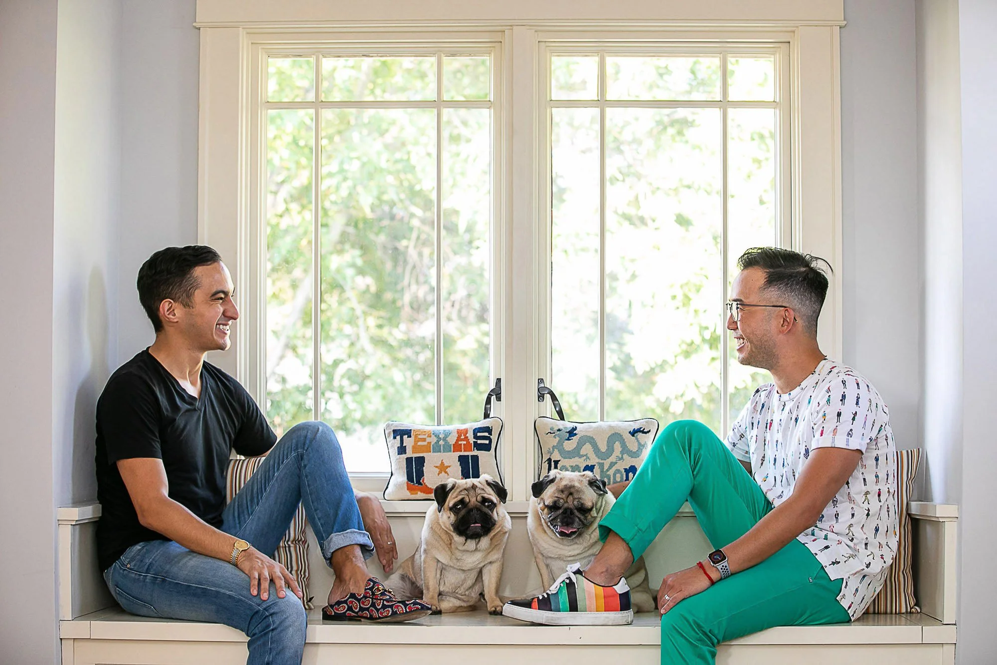 Two men sitting on a window seat, smiling and talking, with two bulldogs between them. The window behind them is bright with greenery outside. The man on the left is wearing a black t-shirt and blue jeans, while the man on the right is wearing glasse