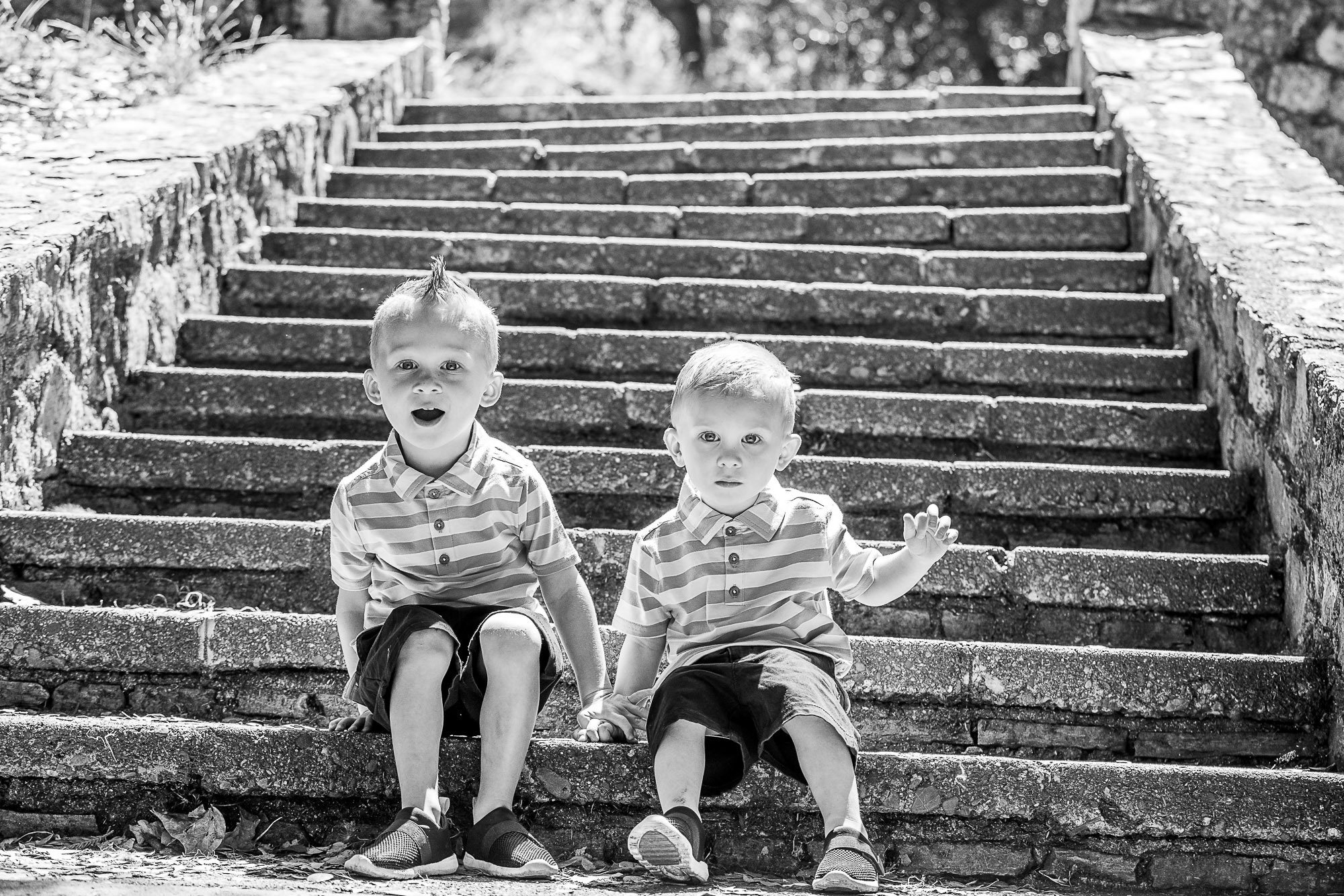 Two young boys sitting on stone steps outdoors, one with a surprised expression and the other waving, both wearing striped shirts and shorts.
