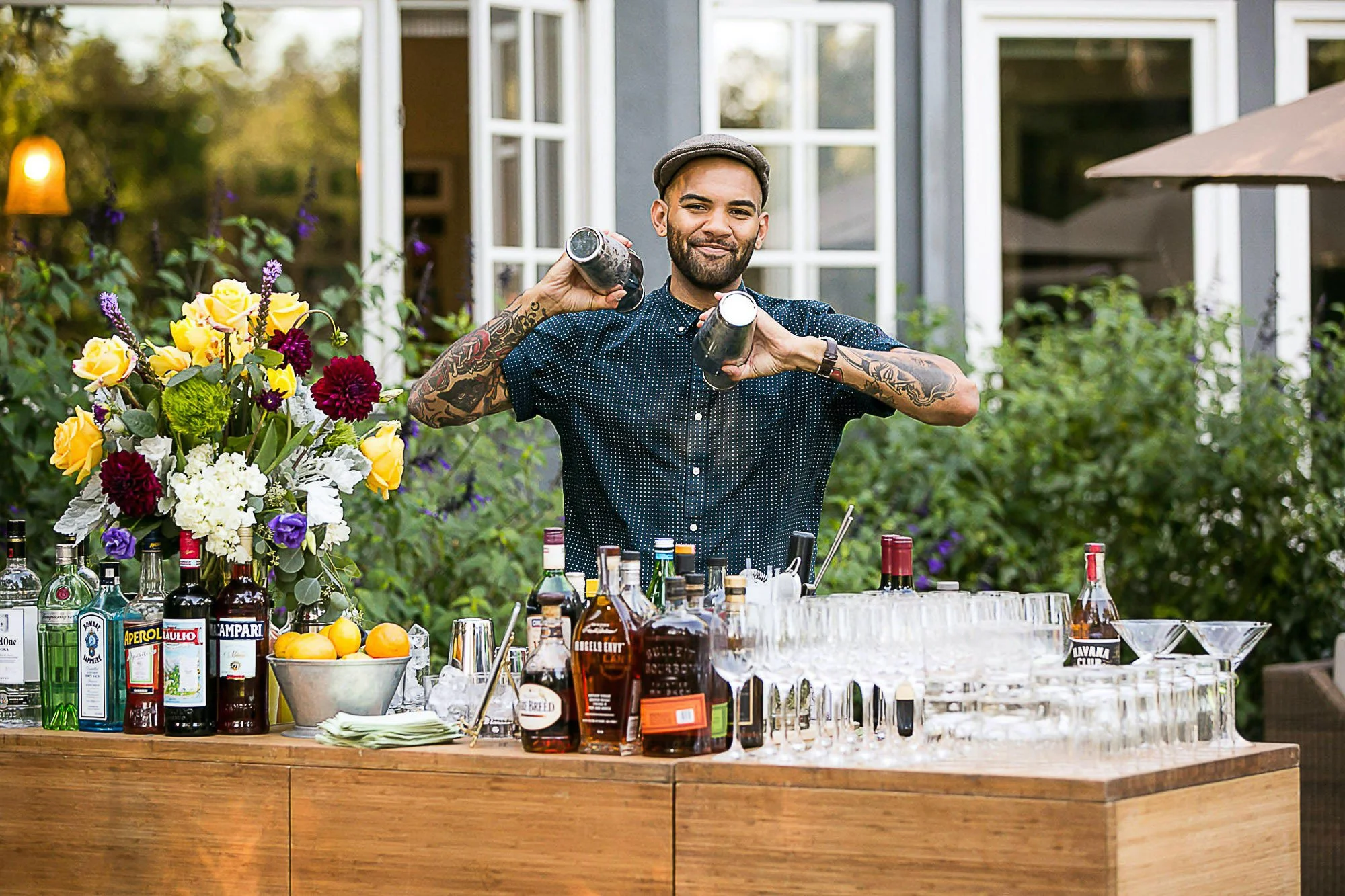 A man with tattoos on his arms and a beard, wearing a gray hat and a navy polka dot shirt, stands behind a bar with various bottles of alcohol and a floral arrangement, holding two cocktail shakers, at an outdoor gathering.