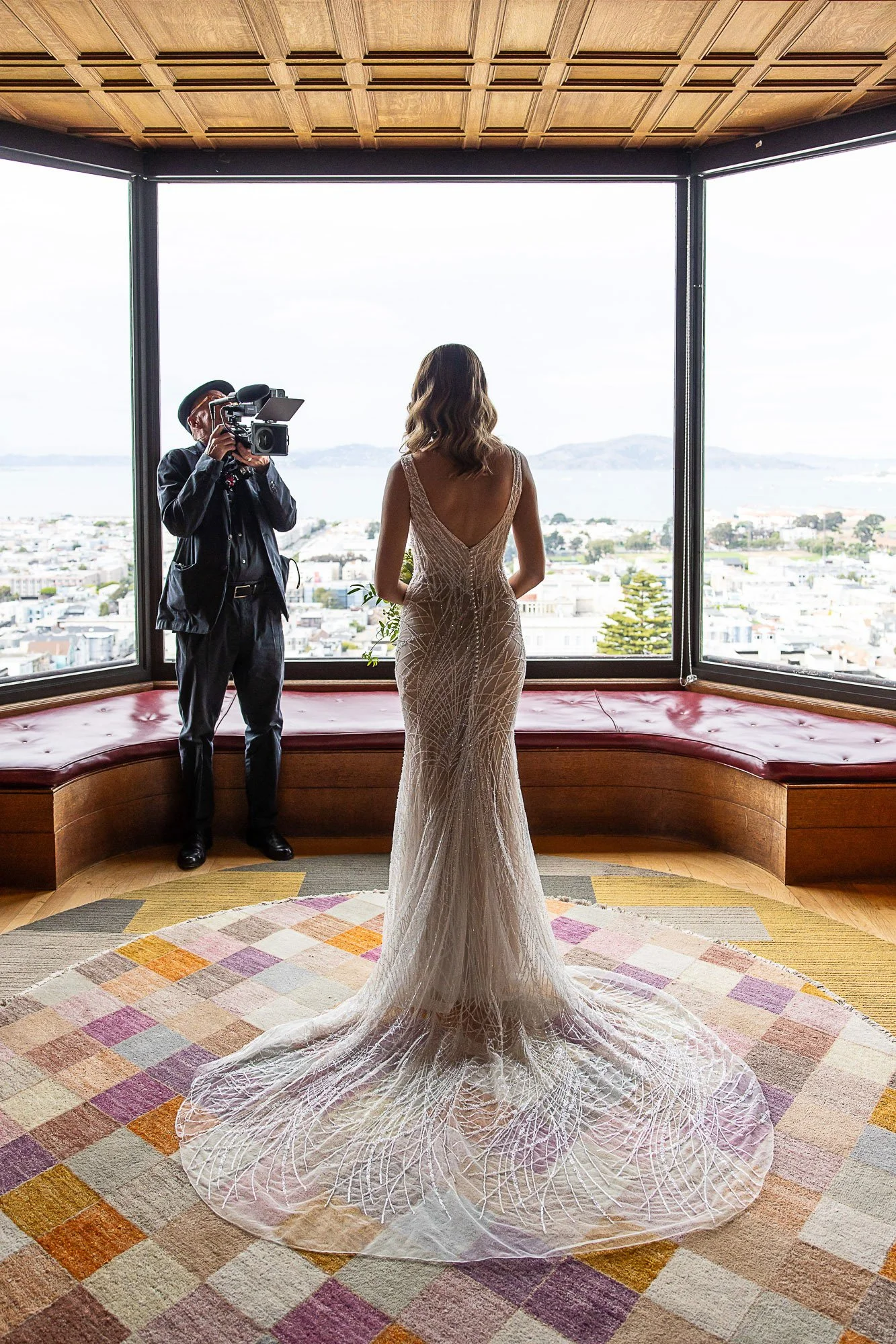 A woman in a white wedding dress with a long train stands inside a room with large windows overlooking a cityscape and water. Arrowood Photography's videographer is filming  her.