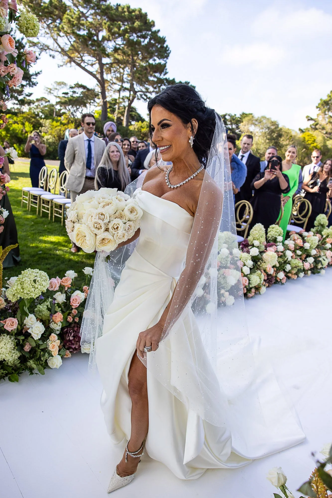 A bride in a strapless white wedding gown holding a bouquet of white roses, smiling, with guests in formal attire in the background during an outdoor wedding ceremony at the Beach and Tennis Club in Pebble Beach surrounded by flowers and greenery.
