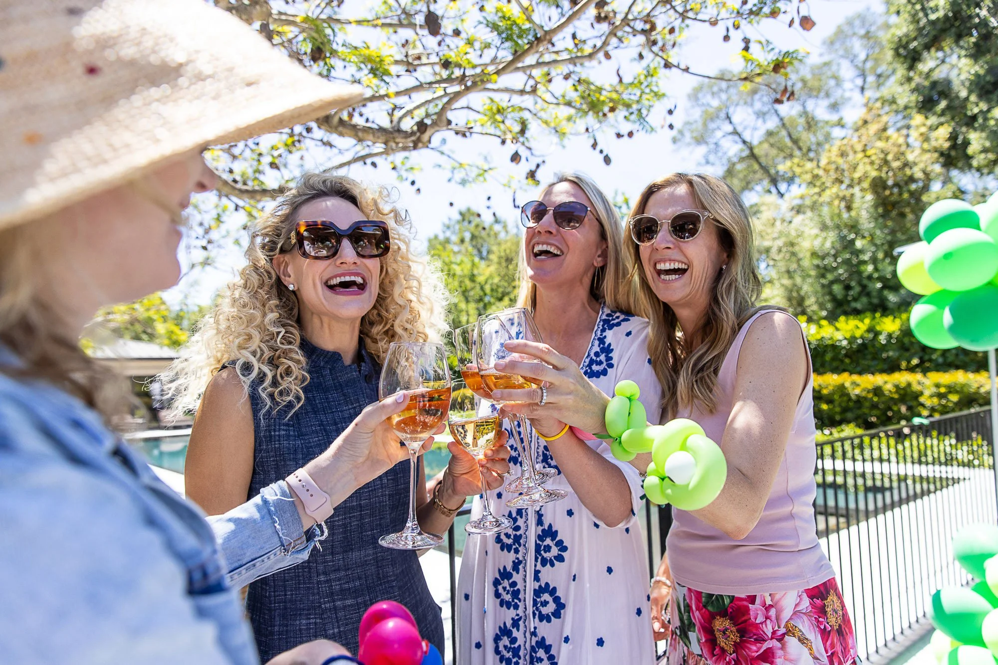 Four women smiling and toasting with wine glasses outdoors at a gathering, surrounded by green trees and balloons.