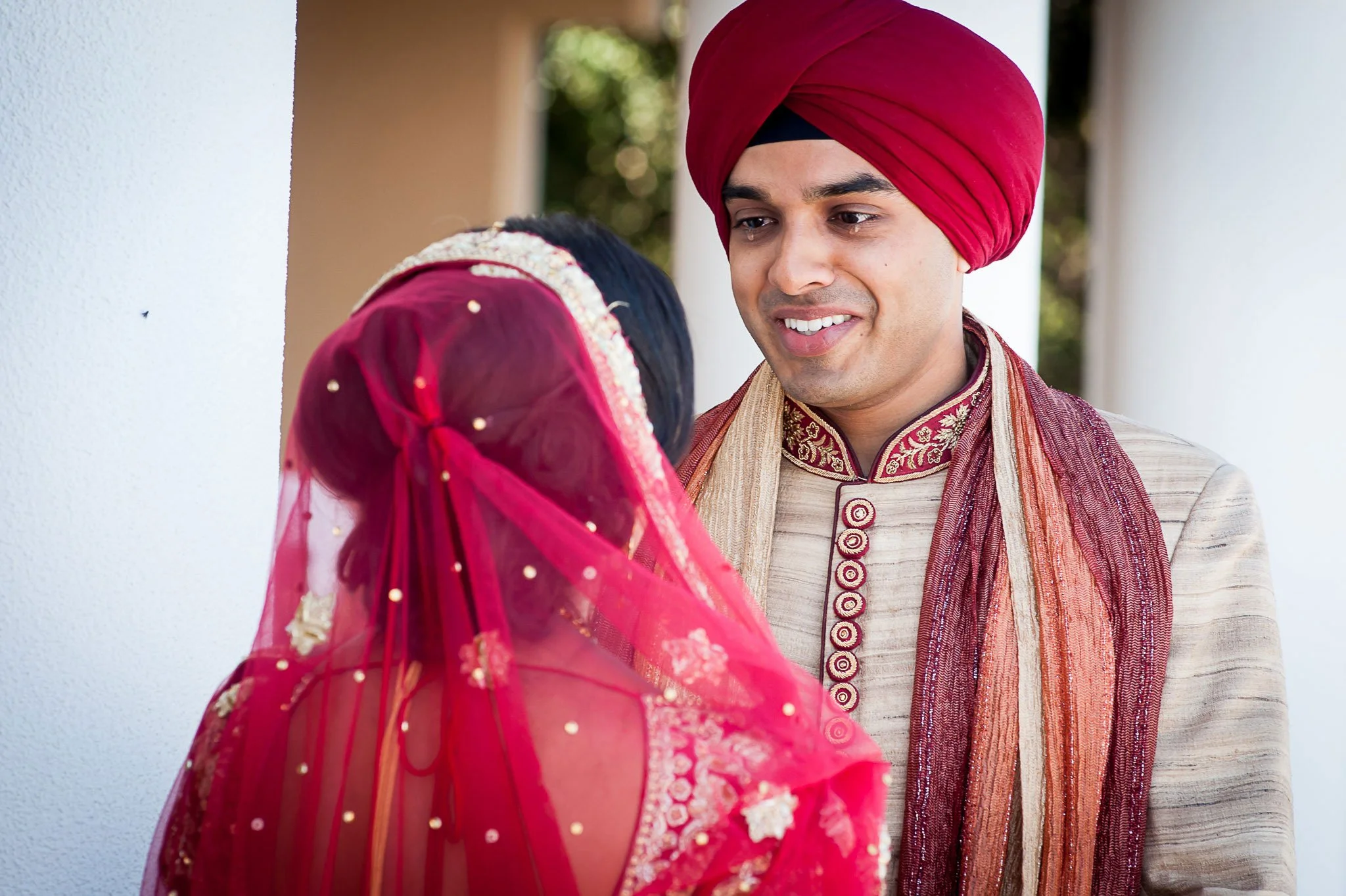 A groom in a cream-colored traditional Indian outfit with red and gold embroidered details, wearing a red turban, smiling and looking at his bride.