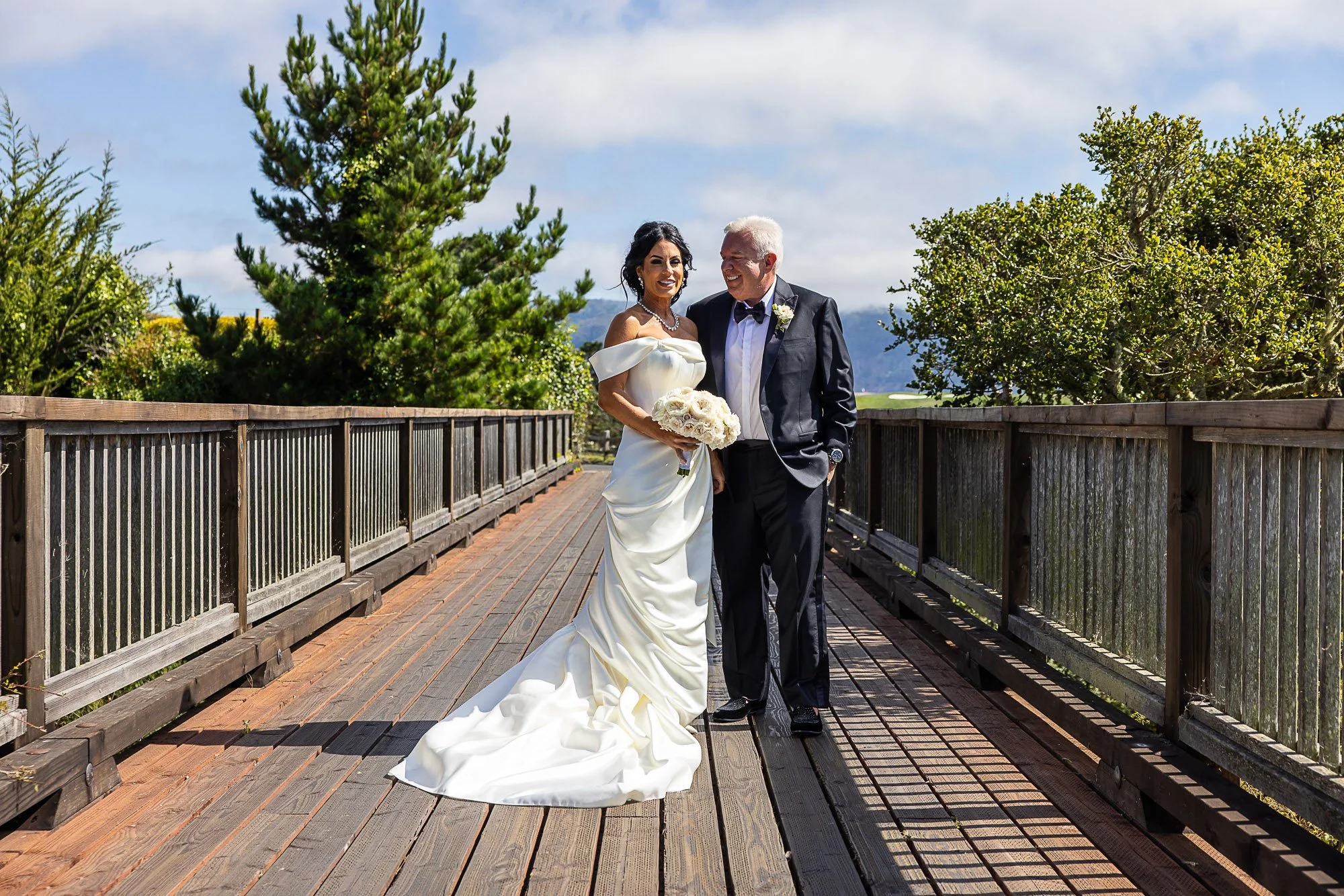 A bride and groom at the Beach and Tennis Club in Pebble Beach, CA walking on a wooden bridge outdoors, smiling and enjoying a sunny day with green trees and blue sky in the background.