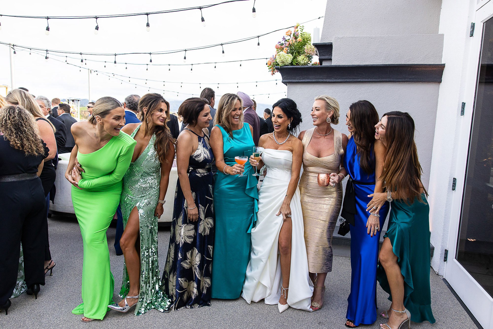 Women in colorful elegant dresses, smiling and talking on a rooftop terrace with string lights, plants, and cityscape in the background at the Beach and Tennis Club in Pebble Beach.