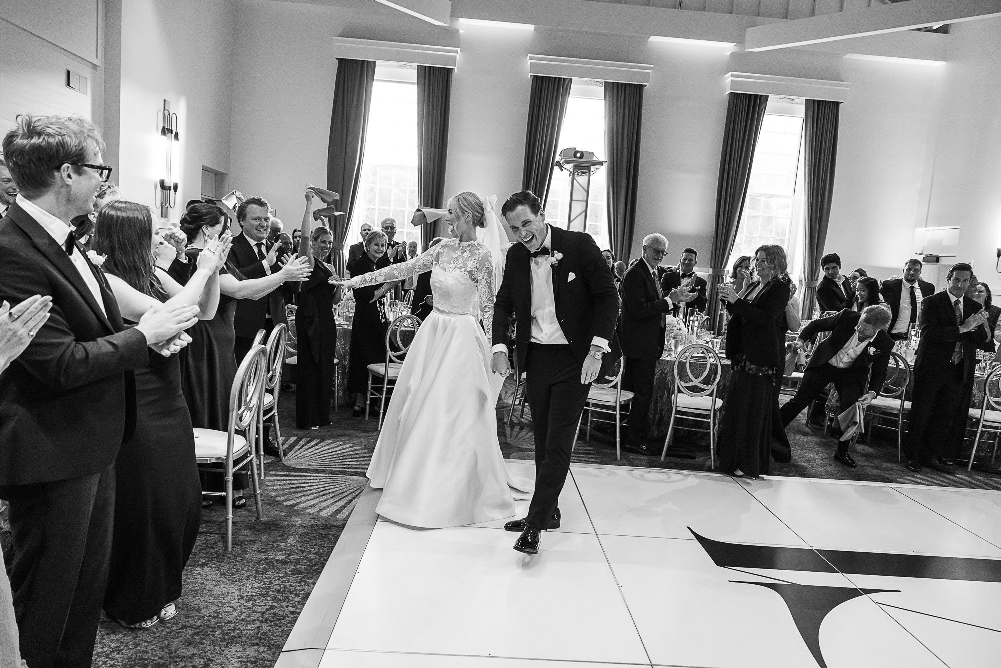 Black and white photo of a bride and groom dancing at a wedding reception, surrounded by guests applauding and smiling inside a decorated banquet hall at Cavallo Point in Sausalito, CA.