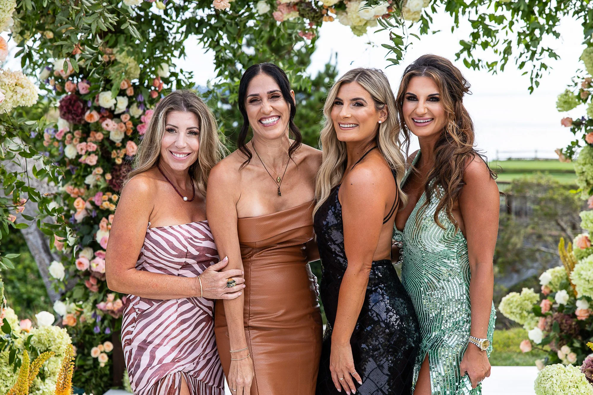 Four women standing together outdoors near a floral arch, smiling for a photo during a daytime event at the Beach and Tennis Club in Pebble Beach, CA.