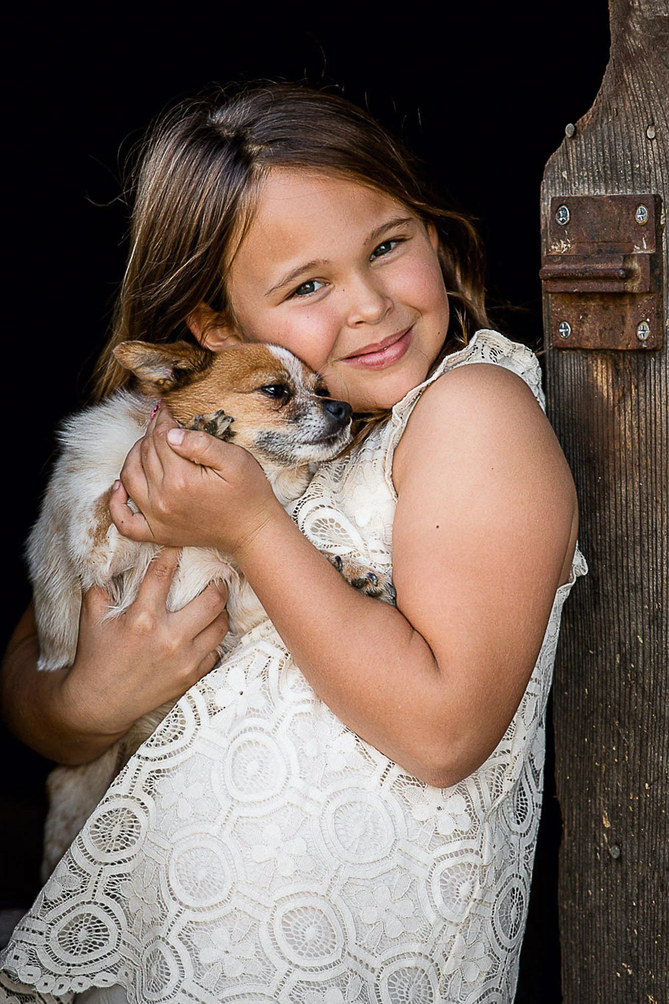 A young girl with light brown hair and blue eyes holding a small brown and white puppy. She is hugging the puppy close to her face, smiling softly. The background is dark, and part of a wooden post is visible on the right side of the image.
