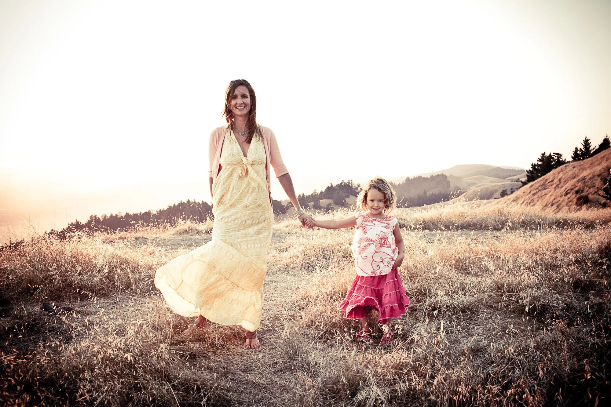 A woman and a young girl walk hand in hand through a grassy field during sunset, with hills and trees in the background.