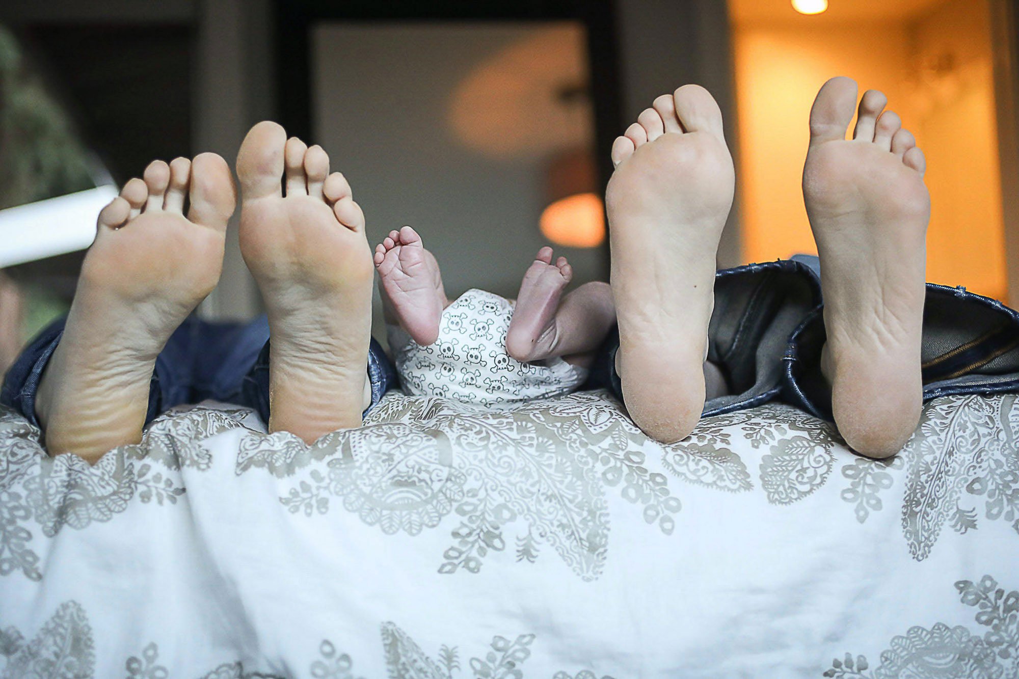 Multiple feet of adults and a baby, all raised in the air, resting on a patterned surface with a background of indoor lighting.