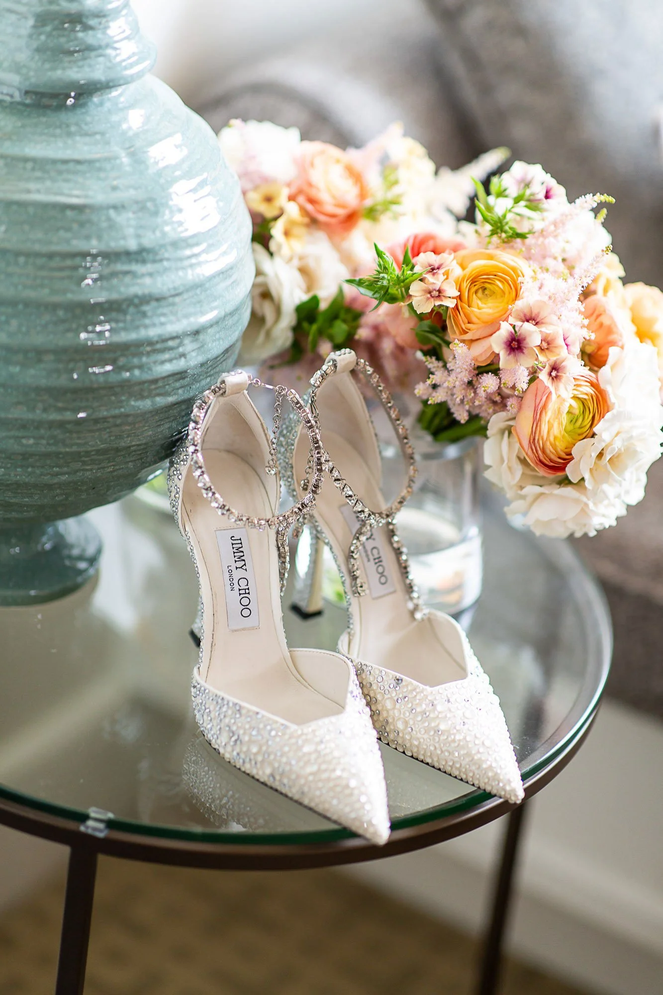 Close-up of a pair of white high-heeled shoes decorated with rhinestones, a bouquet of pink and peach flowers, a large blue vase, and a pair of diamond earrings on a glass table at the Beach and Tennis Club in Pebble Beach, CA.