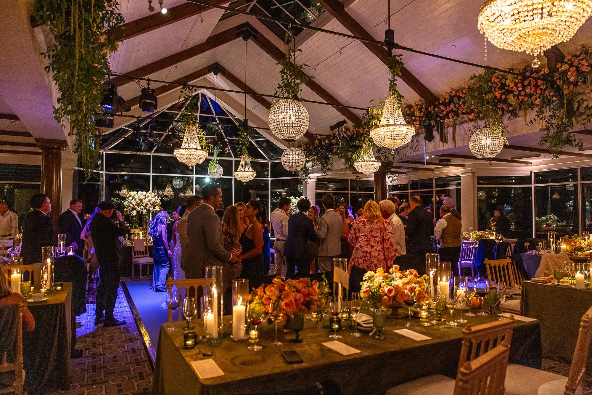 People dressed in formal attire attending a party or wedding reception in a decorated indoor venue with chandeliers, floral arrangements, and candles at the Beach and Tennis Club in Pebble Beach, CA.