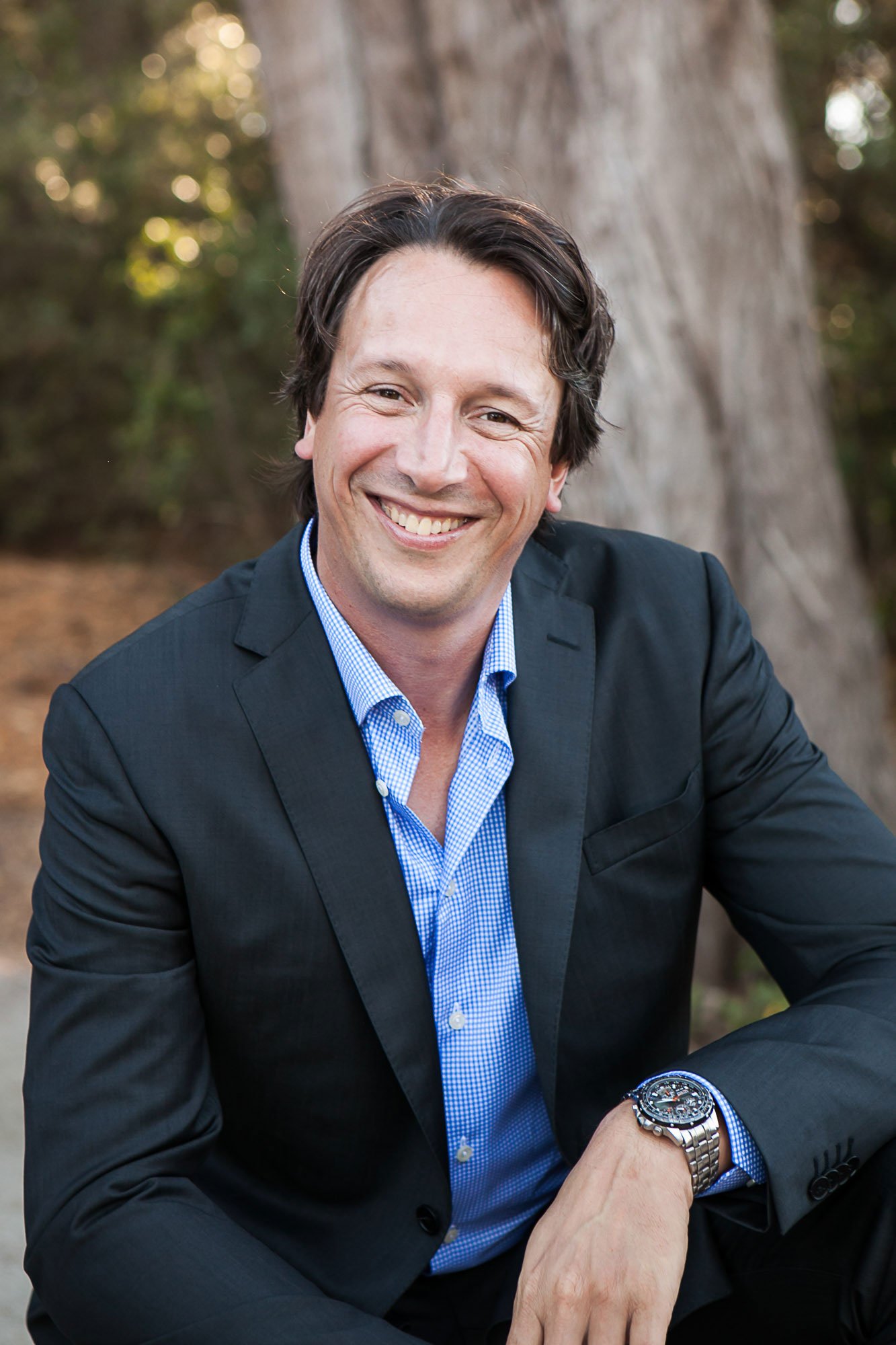 A smiling man in a black blazer and blue checkered shirt, sitting outdoors in front of a large tree, wearing a silver wristwatch.