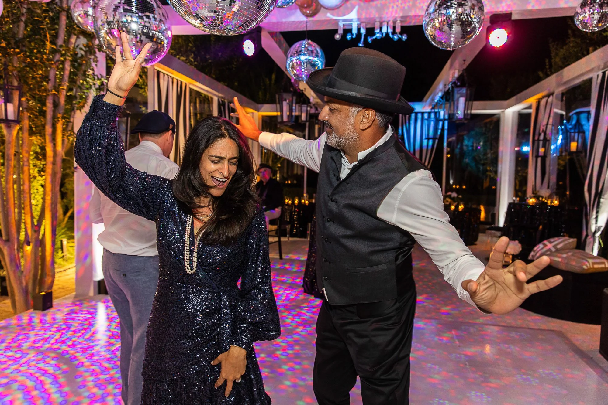 A group of people dancing at a lively party with disco balls and colorful lighting. A man wearing a black hat and vest is dancing with a woman in a dark, glittery dress.