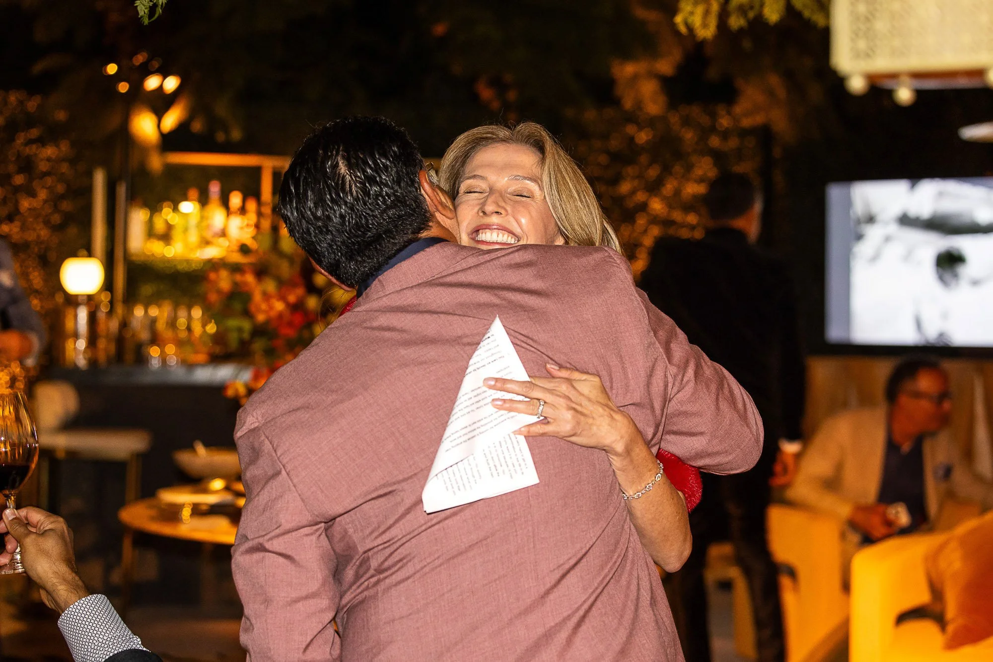 A woman with blonde hair smiling joyfully while hugging a man at a social gathering or celebration in a warmly lit indoor setting.