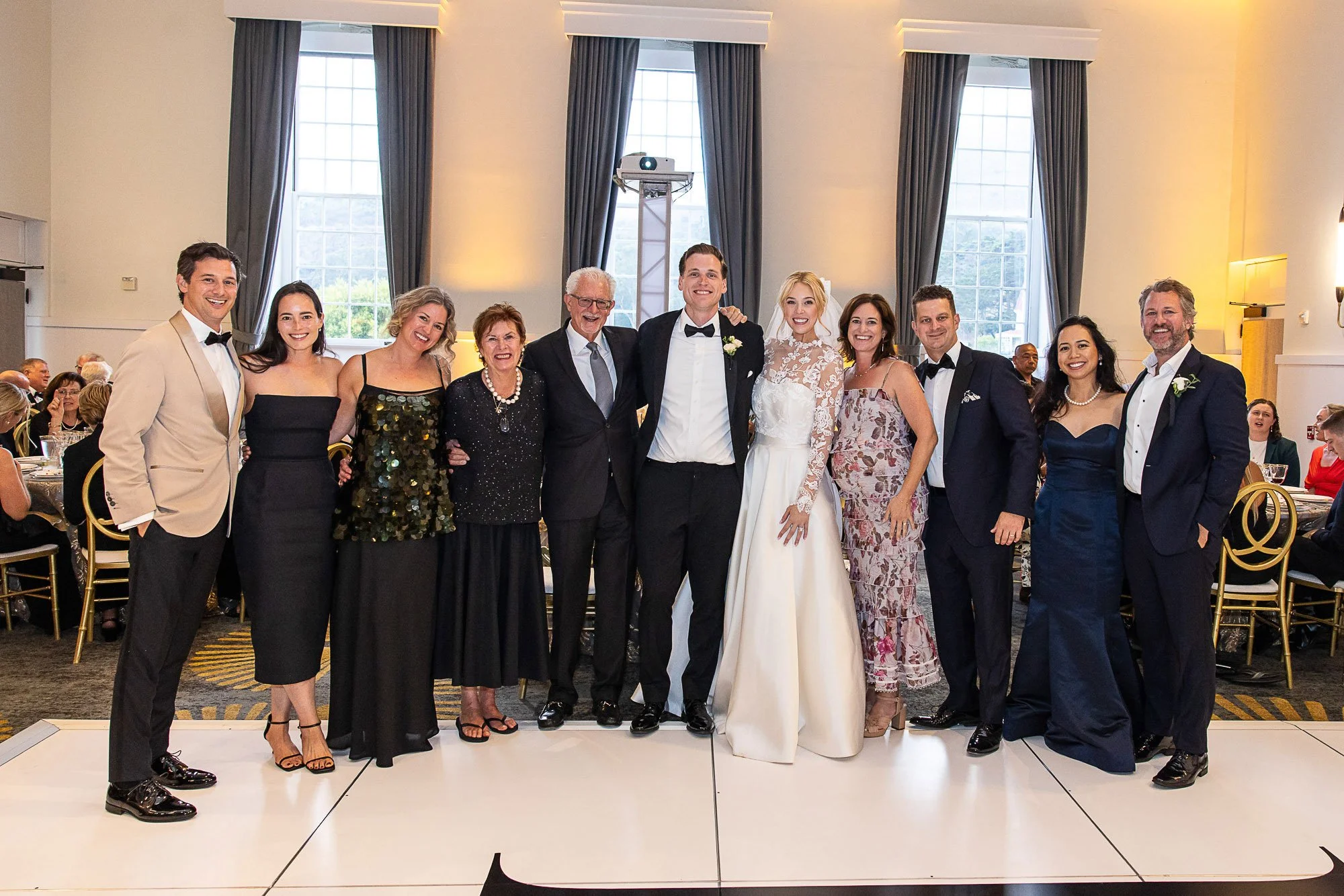 Group of eleven people at a wedding reception, posing for a photo on a raised platform. The bride and groom are in the center, with the bride wearing a white dress with lace sleeves and the groom in a black tuxedo with a bow tie. The wedding party, c